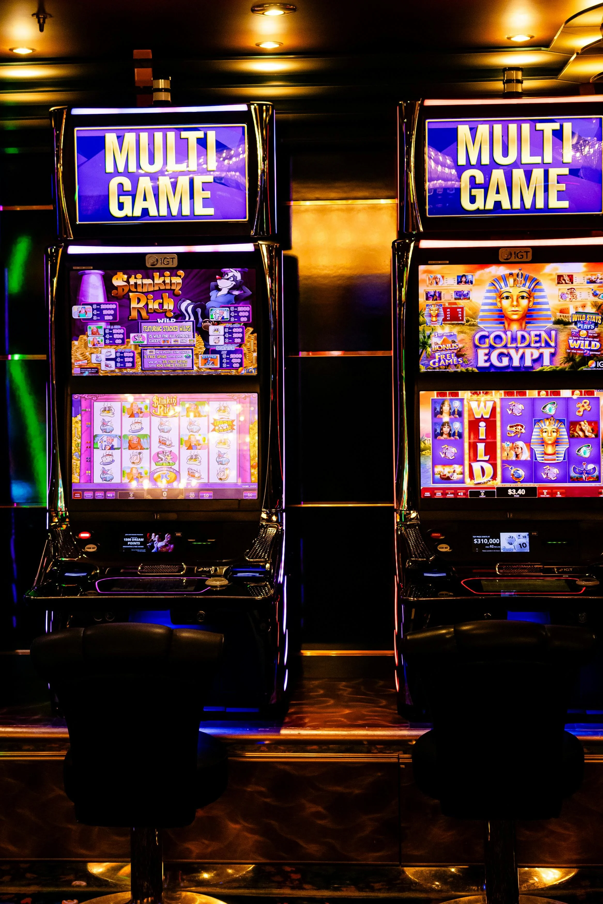 Two brightly lit electronic gaming machines in a casino, both labeled 'MULTI GAME' at the top. The left machine features a game called 'Stinkin' Rich,' and the right machine shows a game named 'Golden Egypt.' There are black chairs in front of each machine.