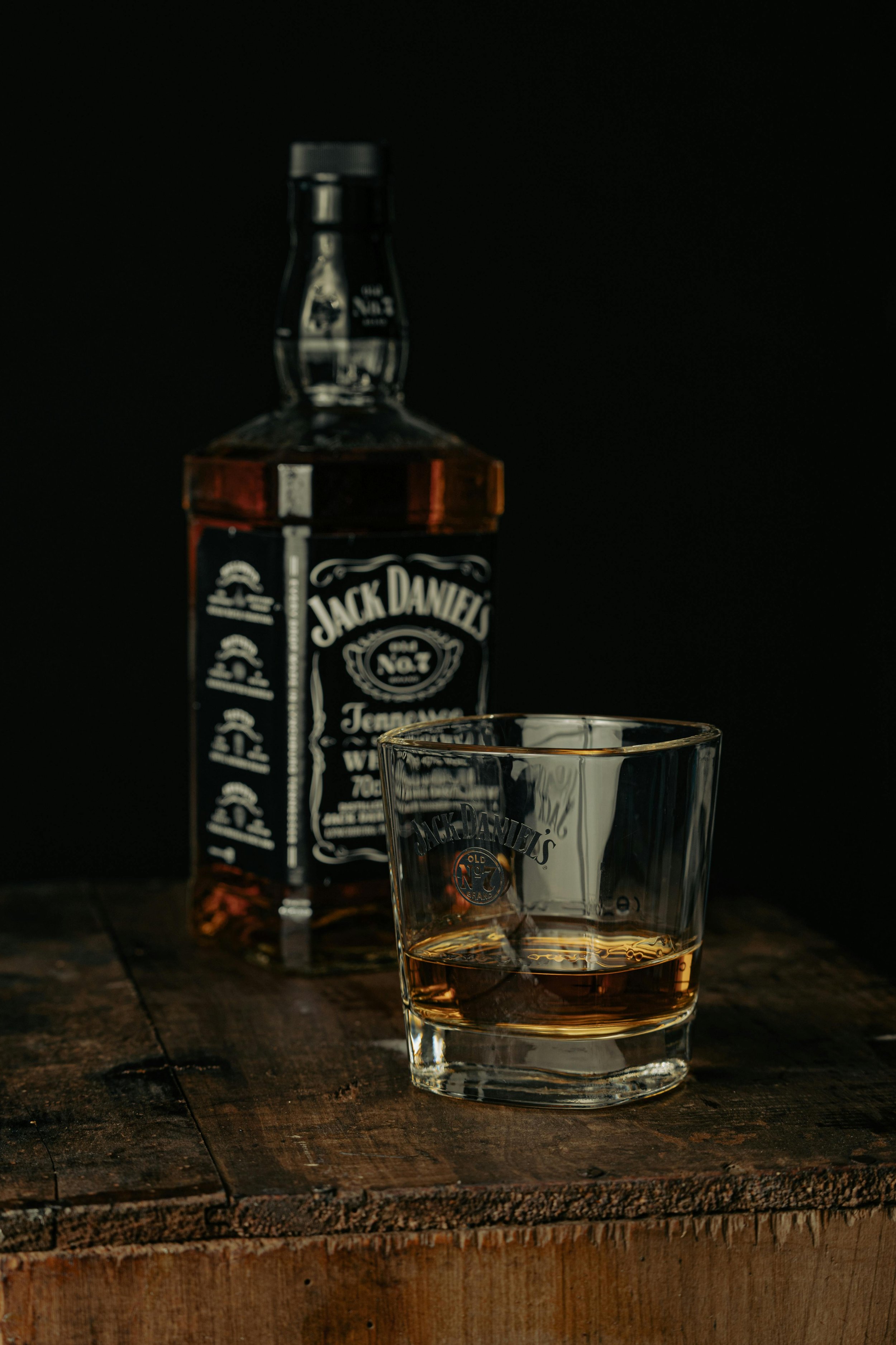 A bottle of Jack Daniel's whiskey and a glass with whiskey on a wooden surface against a dark background.