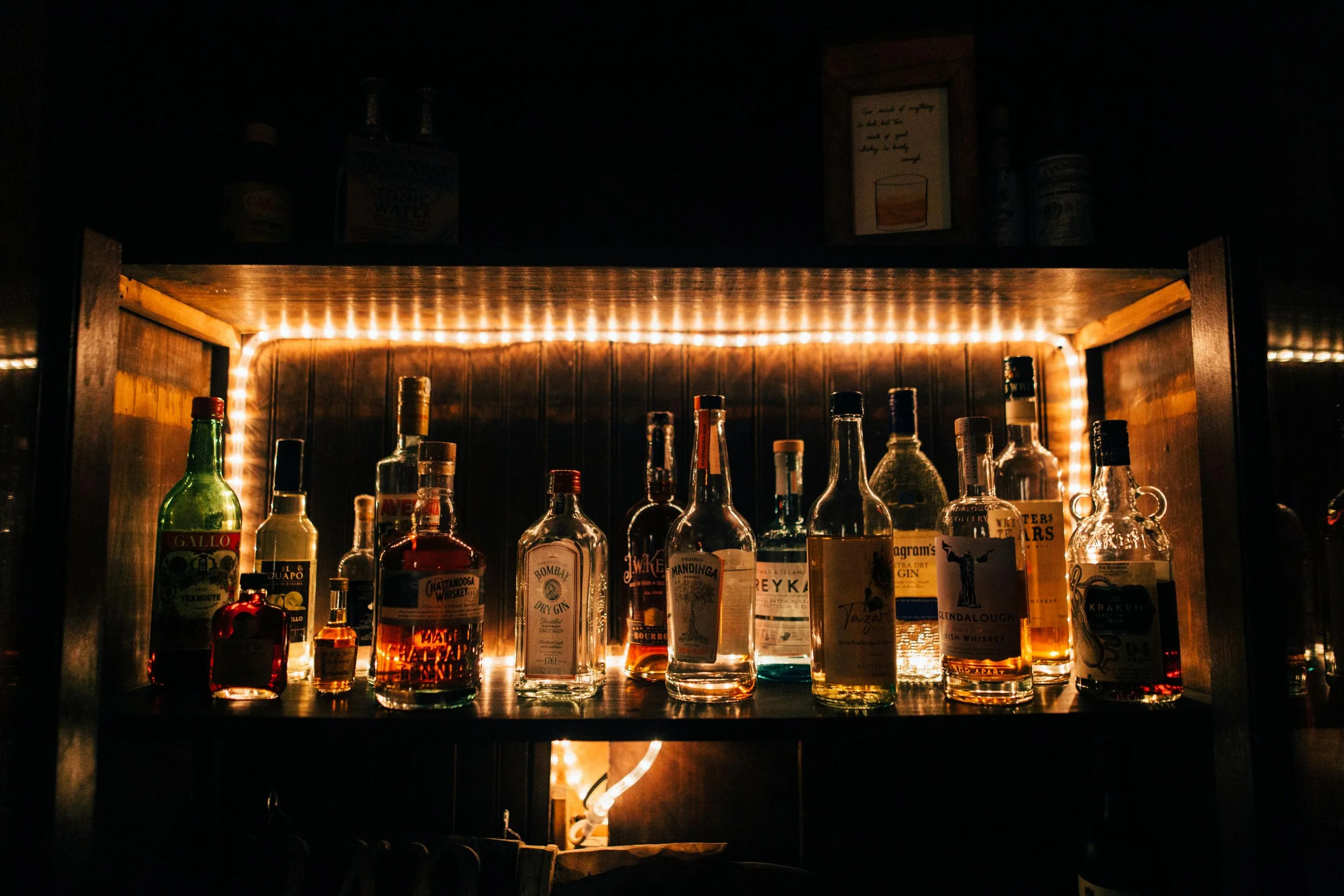 A collection of liquor bottles on a shelf illuminated by warm string lights, in a dark setting.