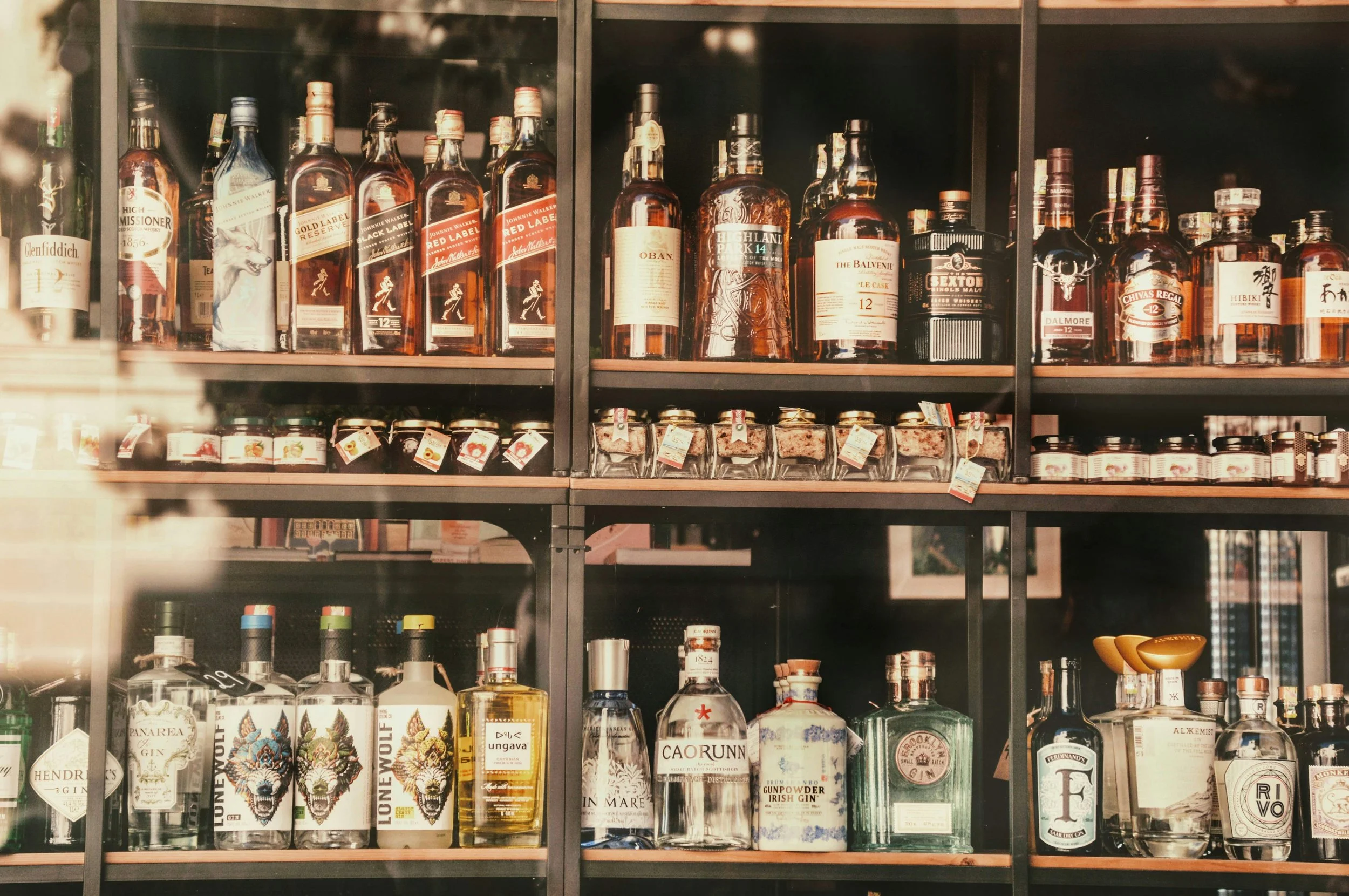 A display of various bottles of whiskey, gin, and other spirits on black shelves, including brands like Glenfiddich, Johnnie Walker, The Balvenie, and others.