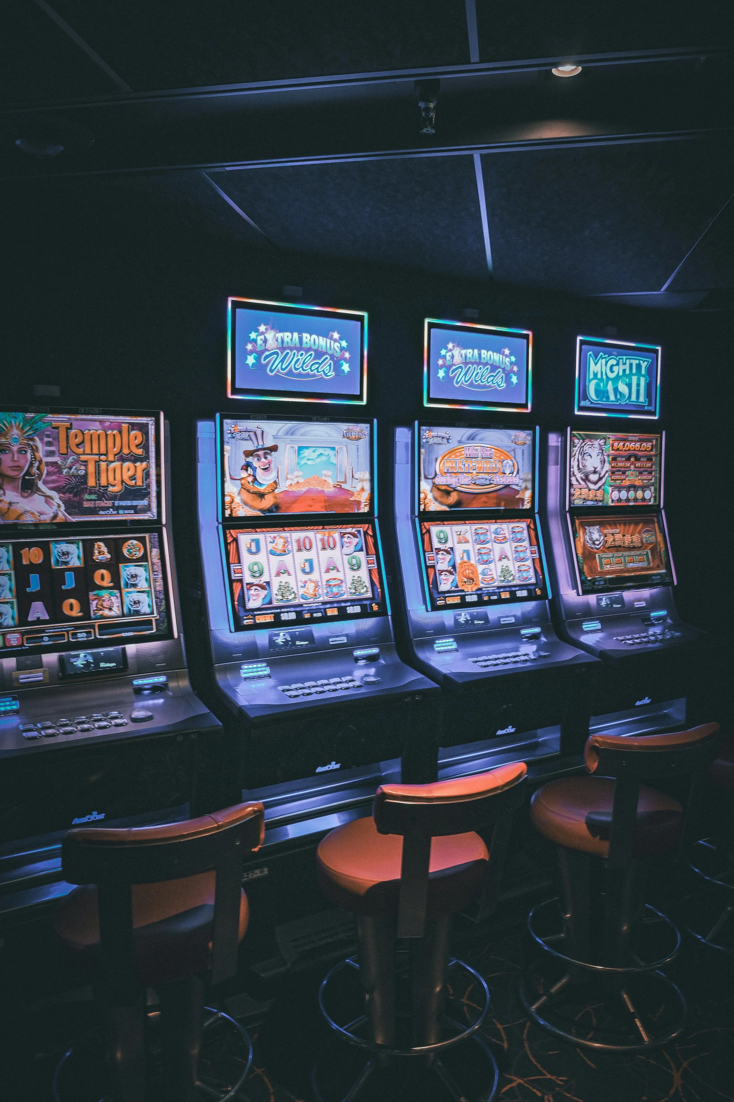Three illuminated slot machines in a dimly lit casino, each with colorful graphics and symbols, and empty chairs in front.