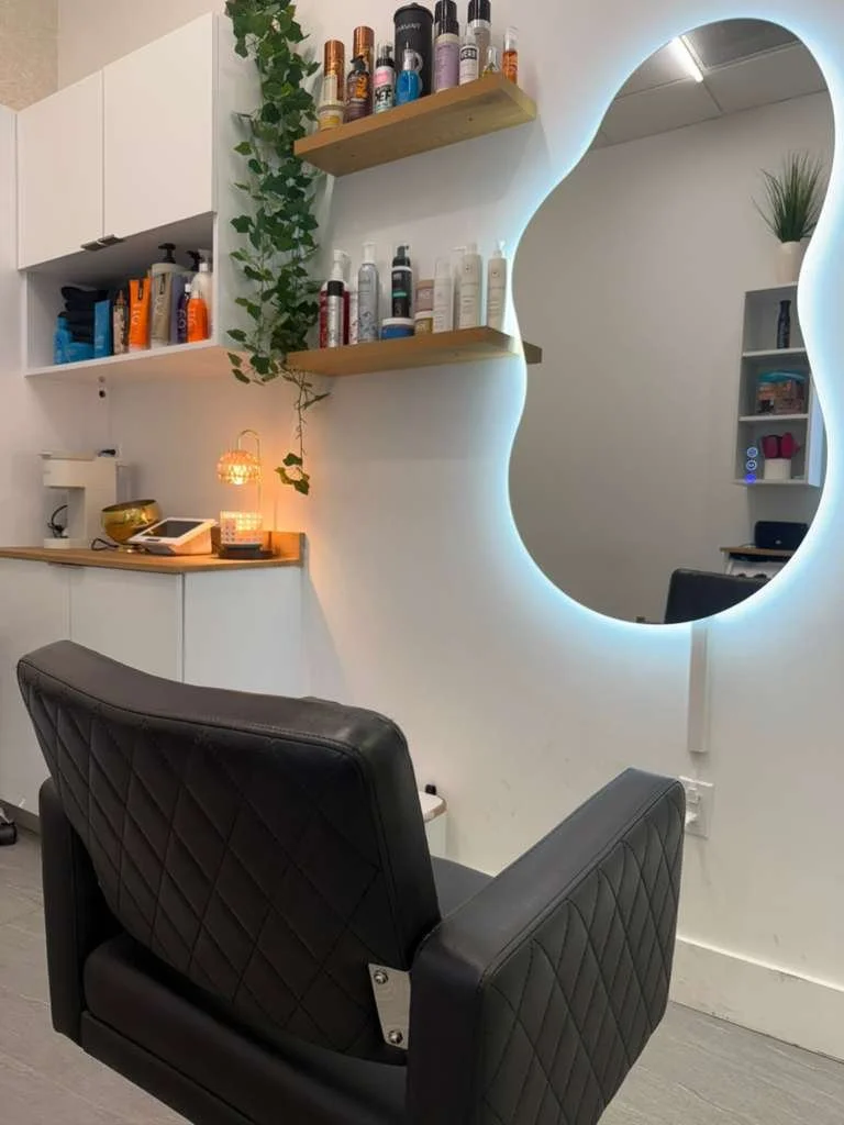 A modern salon or barber shop station with a black leather chair, illuminated mirror, white wall with wooden shelves holding bottles, a potted plant, and various hair and beauty products.