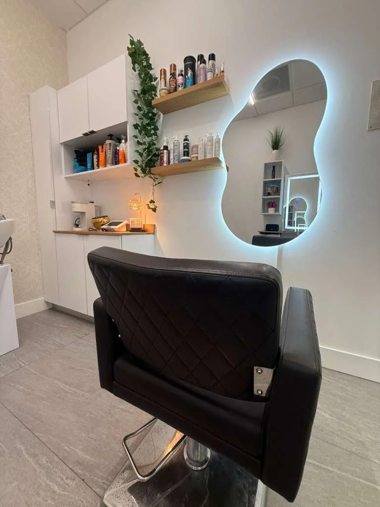 A hair salon with a black salon chair facing a large mirror with a wavy illuminated border. Behind the chair, there are white cabinets and wooden shelves holding hair products, a small lamp, and decorative plants.
