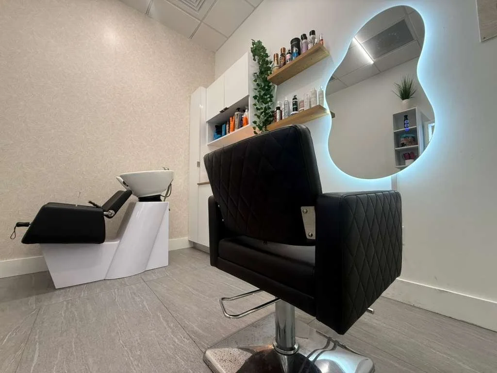 A modern salon interior with a black salon chair and a white washing basin. Shelves with hair products and a plant are on the wall, and a decorative mirror with backlit LED lighting is visible.