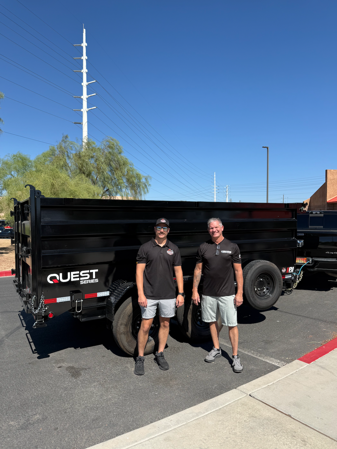 Two men standing next to a black Quest Series trailer in a parking lot on a bright, sunny day. One man is wearing a black shirt and gray shorts, and the other is wearing a black shirt and white shorts. They are smiling at the camera with a clear blue sky and power lines in the background.