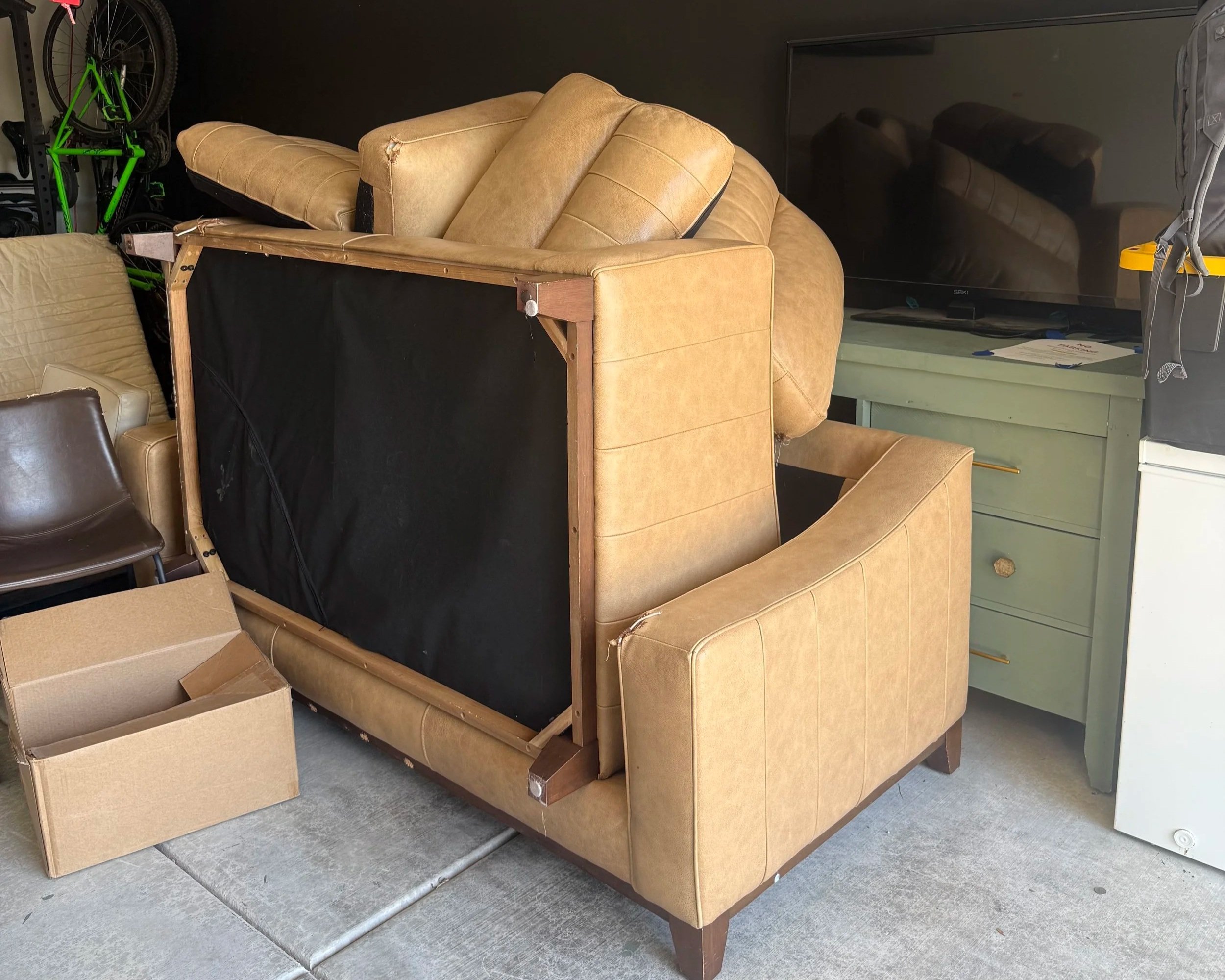A partially assembled beige couch with a black fabric backing, a matching beige armchair, and several cardboard boxes in a room with a gray carpeted floor and a black wall.