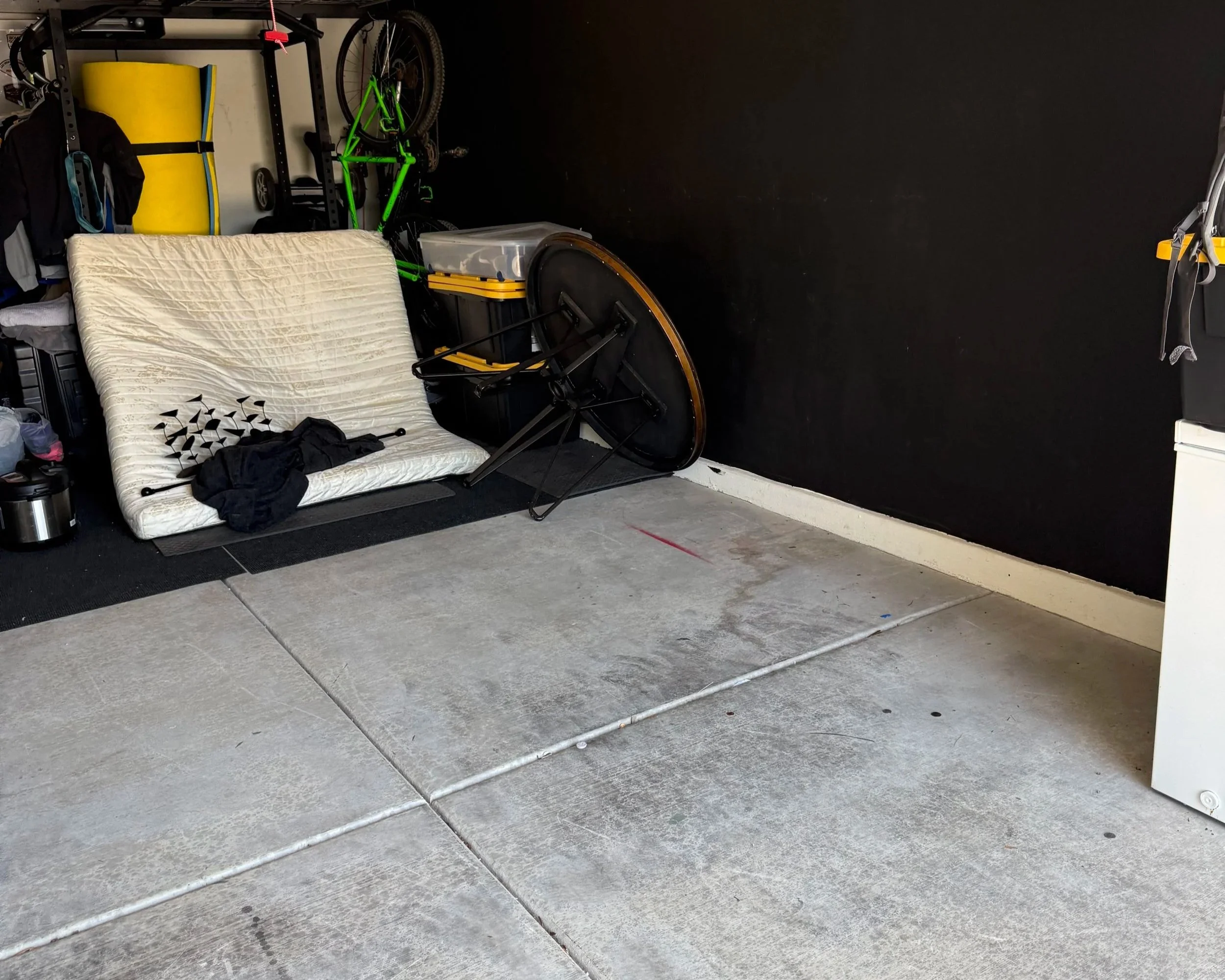 A cluttered garage space with a black wall, a mattress leaning against the wall, a green bicycle, a yellow storage bin, a clear plastic container, a folded round table, and various tools and items scattered on the floor and shelves. The floor has concrete tiles with a metal pipe running along the edge.