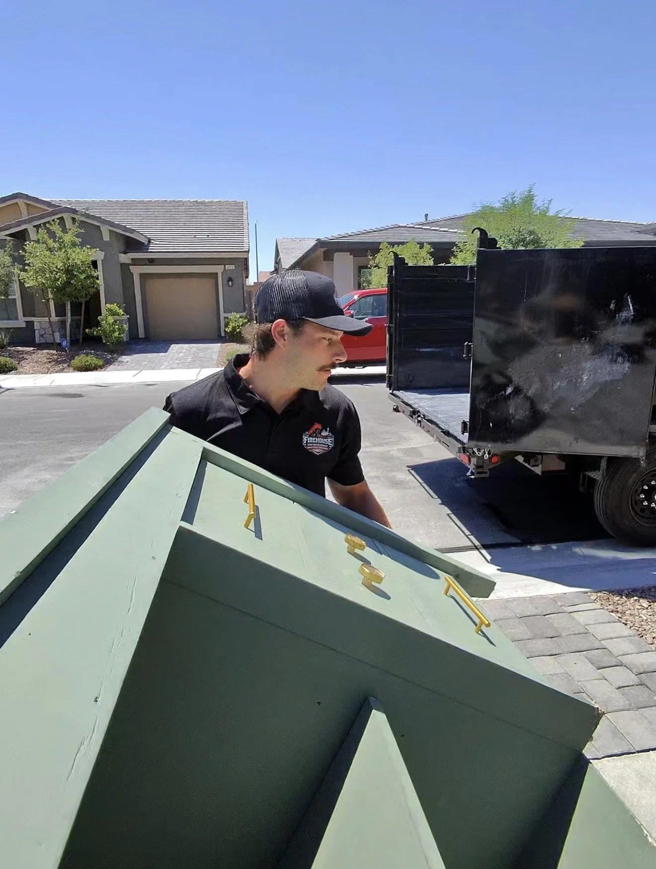 A man with a mustache and black cap loading a green shed into a truck on a suburban street with houses and trees under a clear blue sky.