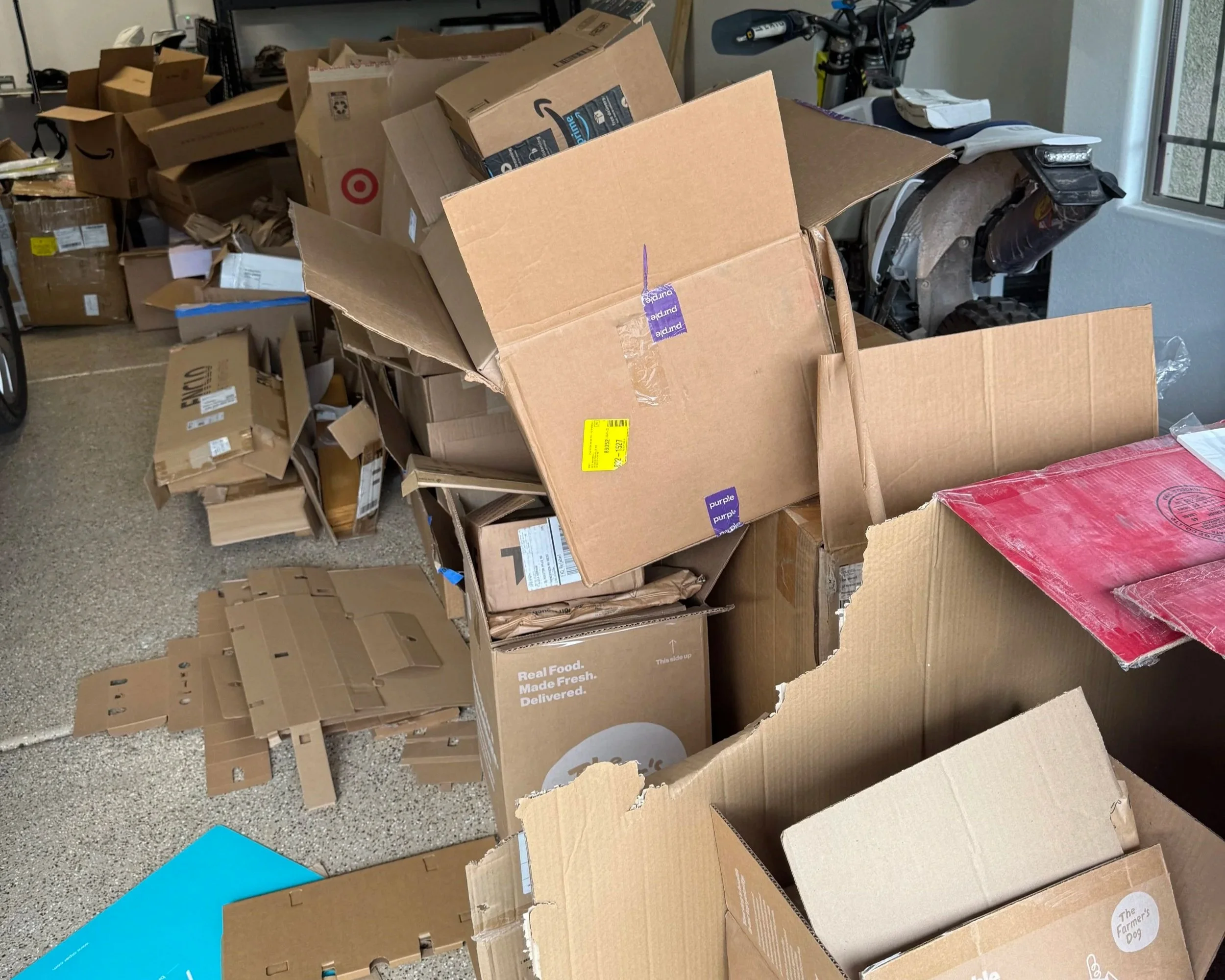 A cluttered garage with many open and unopened cardboard boxes, some flattened, along with a parked motorcycle near a window.