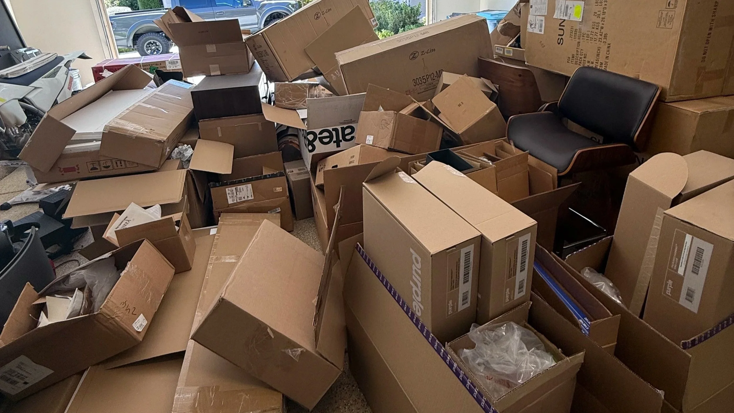 A room filled with numerous cardboard boxes, some open and some closed, with packing materials and labels, next to a large window with a view of parked cars outside.
