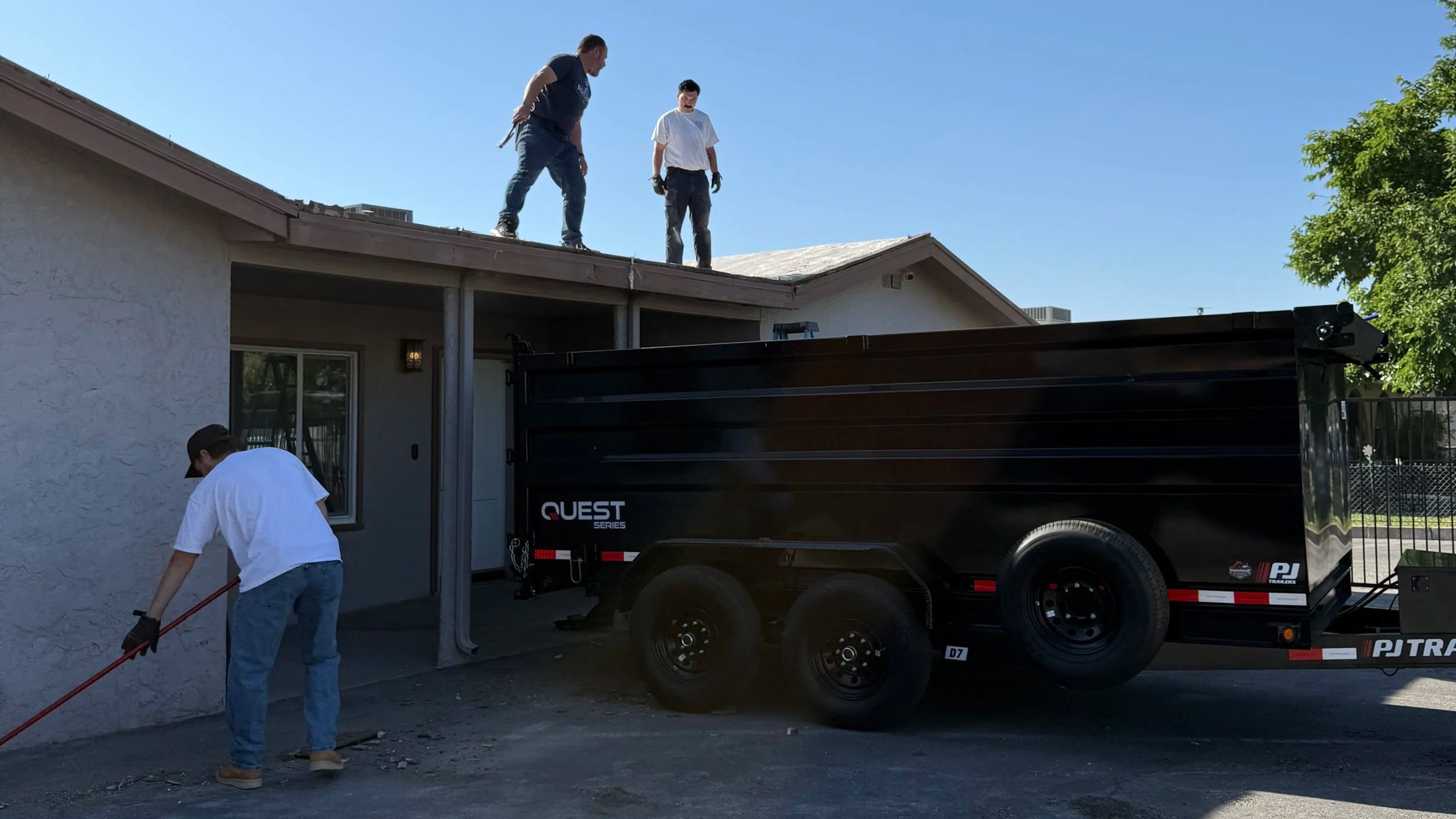 Two men on the roof of a house working on repairs, with one man on the ground holding a tool, and a trailer parked in front of the house on a sunny day.
