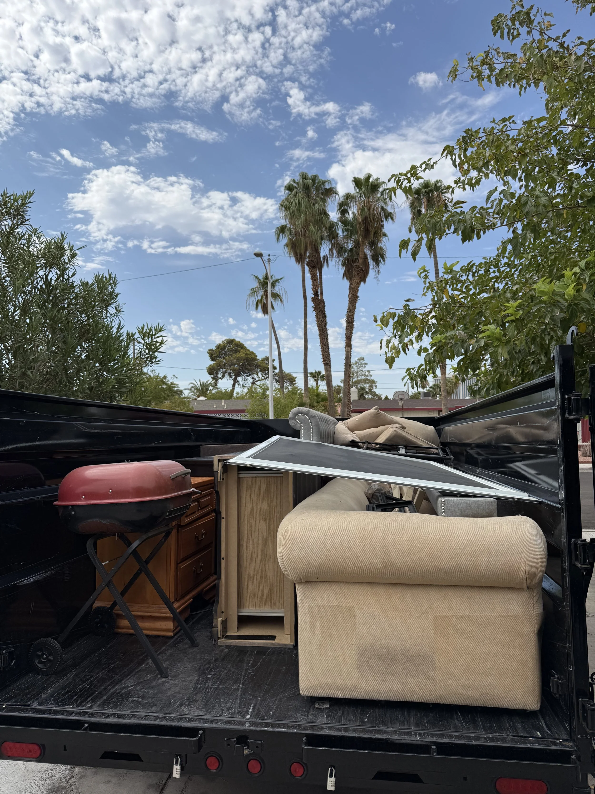 View from the back of a truck filled with furniture and household items, with palm trees and a partly cloudy sky in the background.