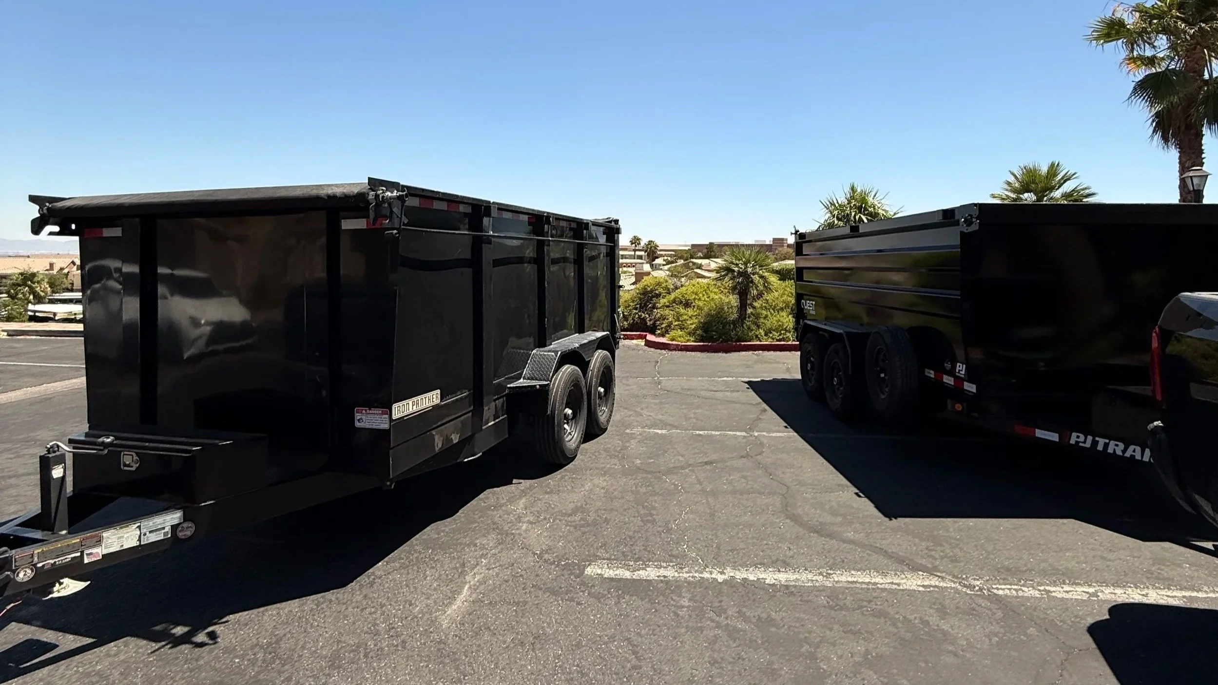 Two black trailers parked in a lot with trees and buildings in the background under a clear blue sky.
