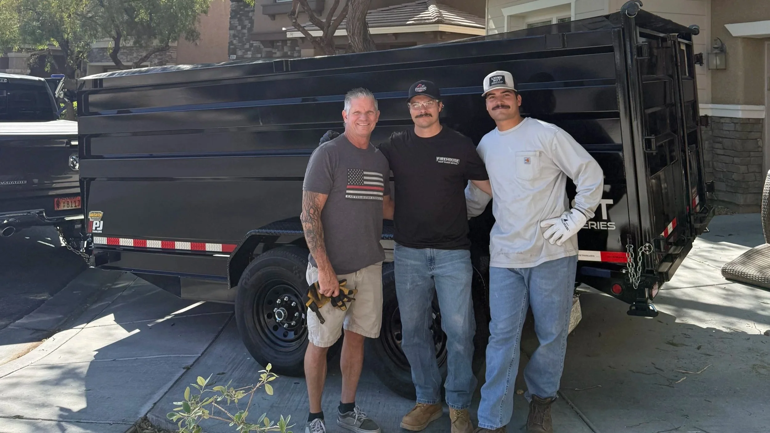 Three men standing outdoors in front of a black utility trailer, smiling. The man on the left has tattoos on his right arm, is wearing a gray T-shirt with an American flag, and light-colored shorts. The middle man has dark hair and a mustache, is wearing glasses, a black T-shirt, and jeans. The man on the right has a mustache, is wearing a white long-sleeve shirt, jeans, and a cap. There are residential houses and trees in the background.
