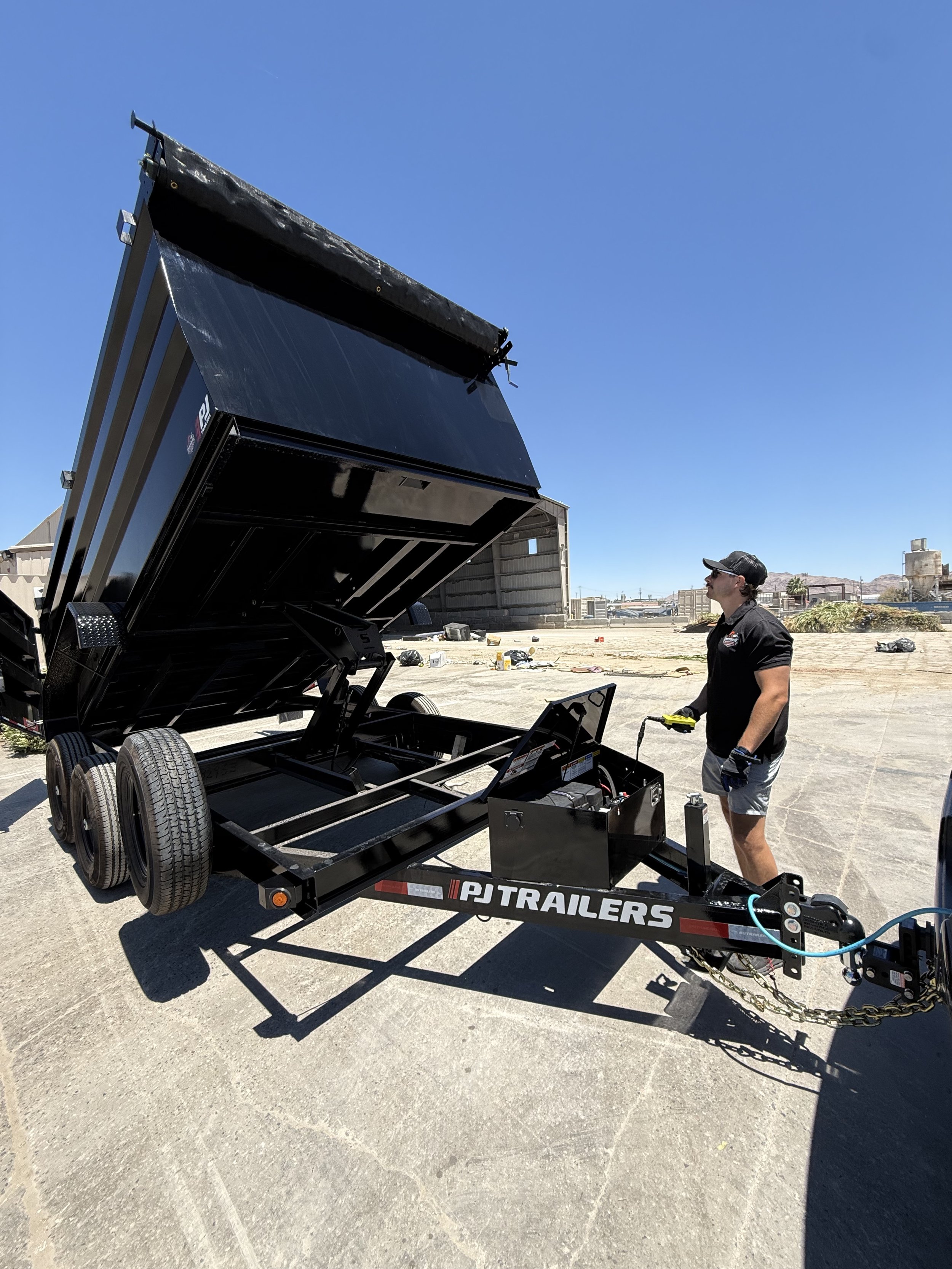 A man wearing sunglasses, a baseball cap, black gloves, and a dark shirt stands next to a black trailer with an open raised bed, in an outdoor area with clear blue skies and mountains in the background. The trailer has the word "TRAILERS" written on the side.