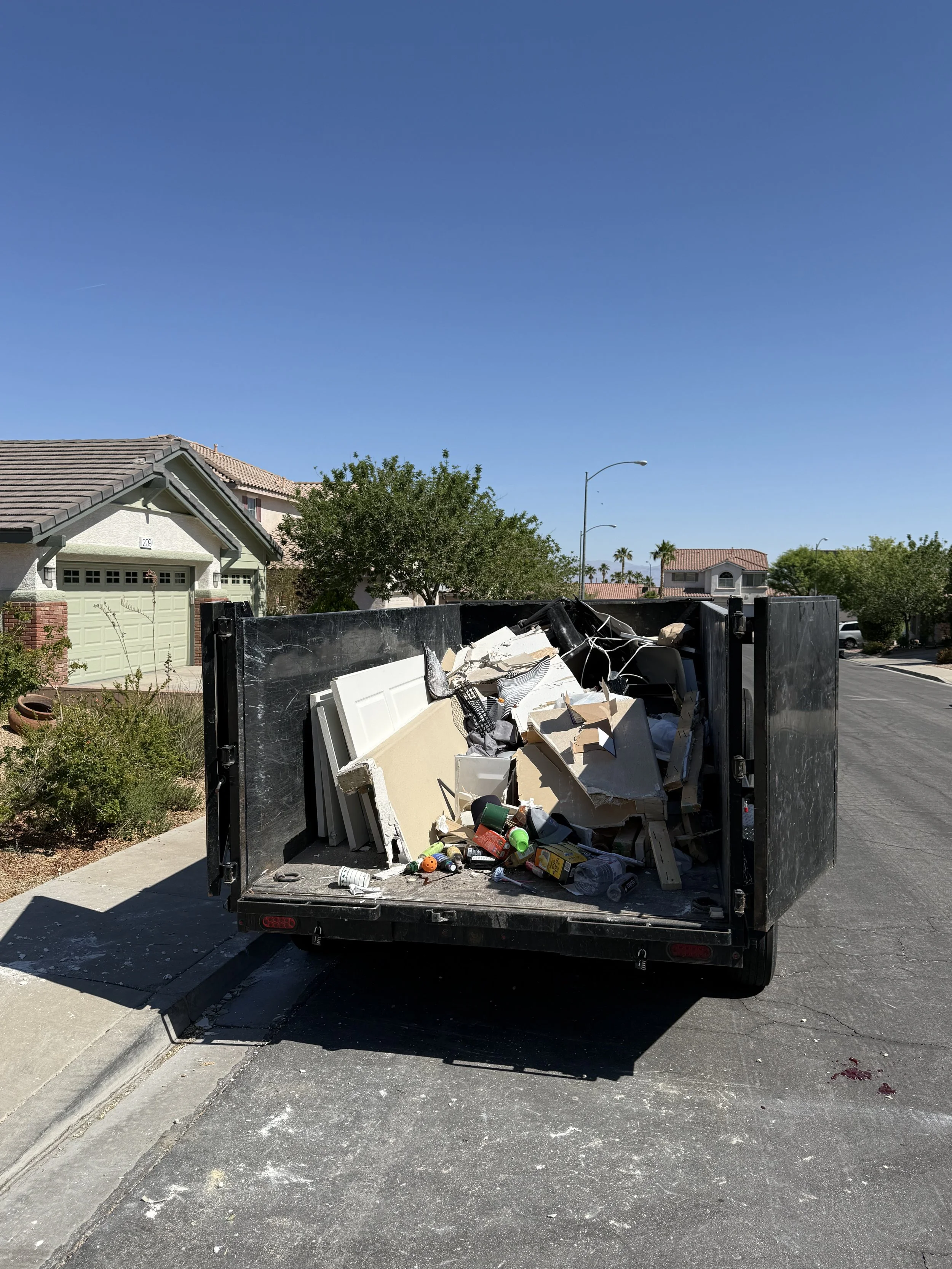 Dump truck filled with debris, trash, and household items parked on a residential neighborhood street under clear blue sky.