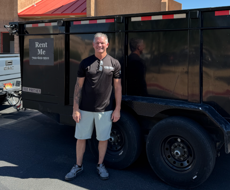 Man standing in front of a black trailer with a sign that reads 'Rent Me' and a phone number, in a parking lot.