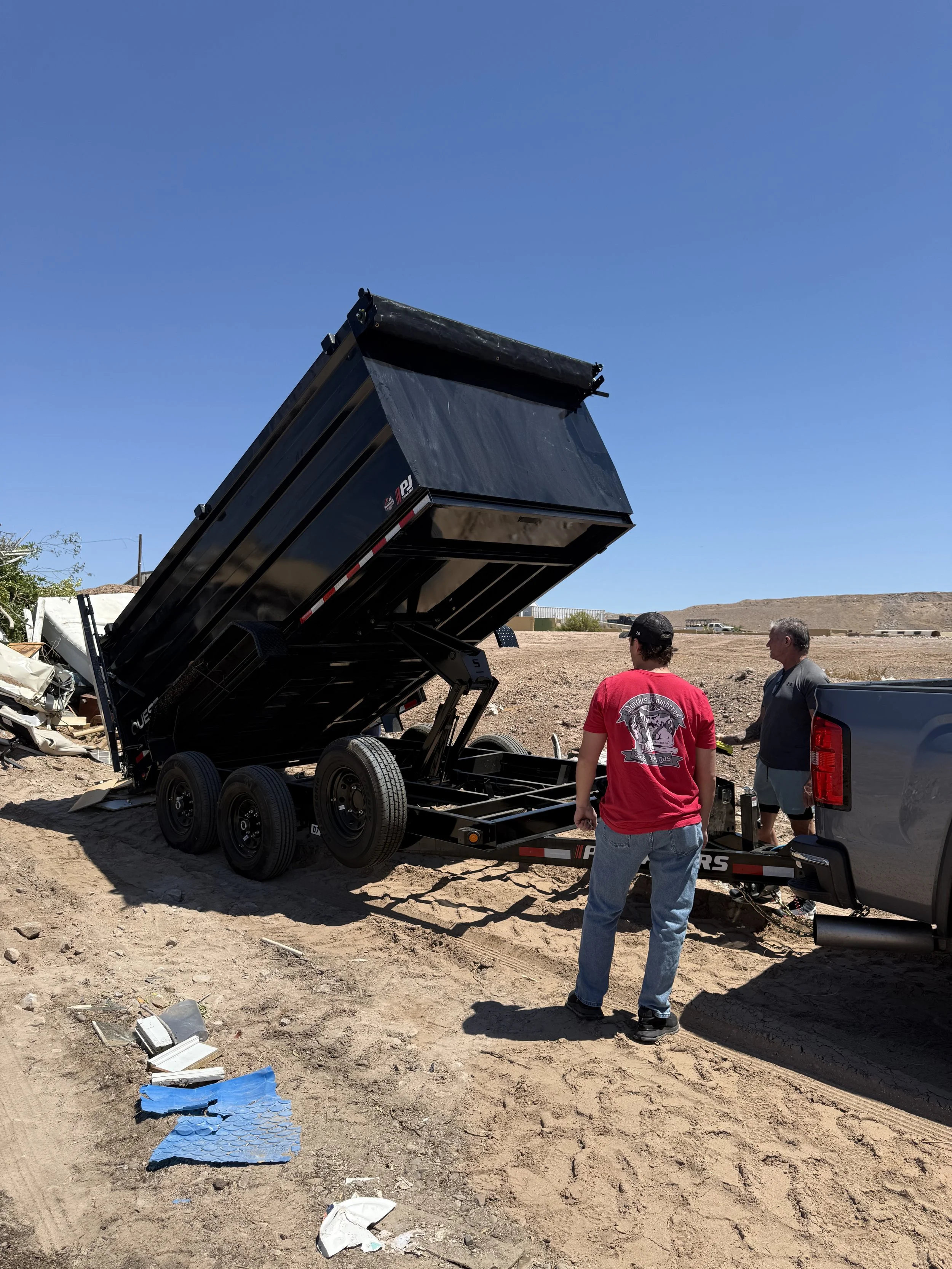 Two men standing near a black dump trailer with the bed raised on a dirt lot. Debris and trash are scattered on the ground, and a truck is nearby. The sky is clear and blue, with some distant hills in the background.