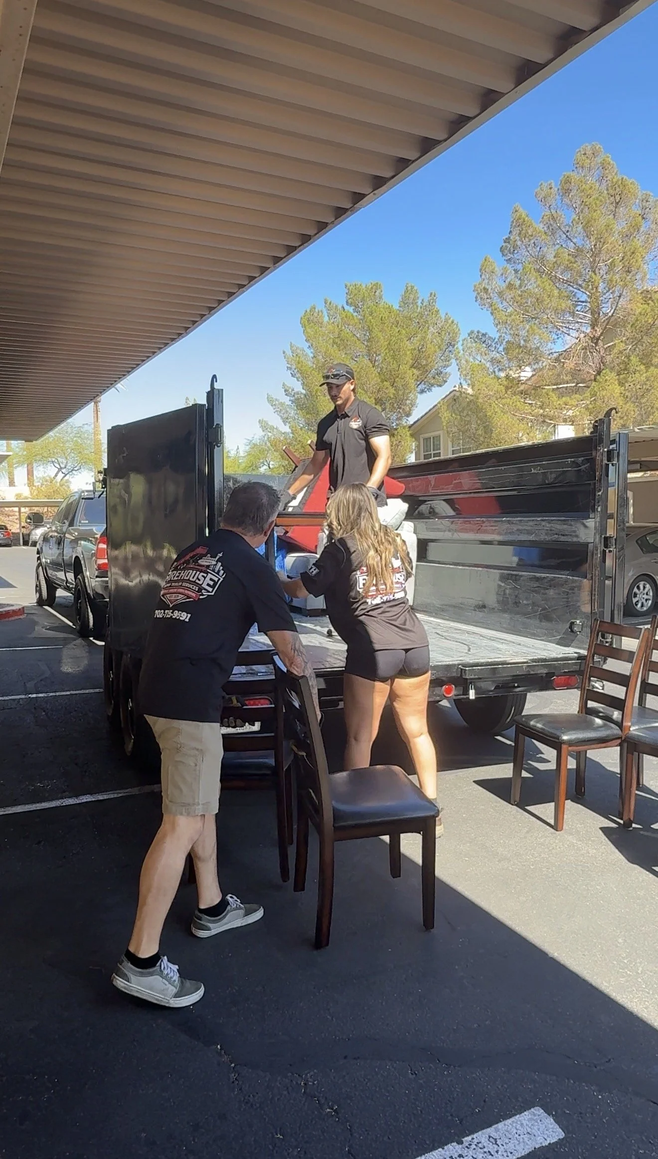 People unloading a motorcycle from a black flatbed trailer in a parking lot under a shaded roof, with trees and cars in the background.