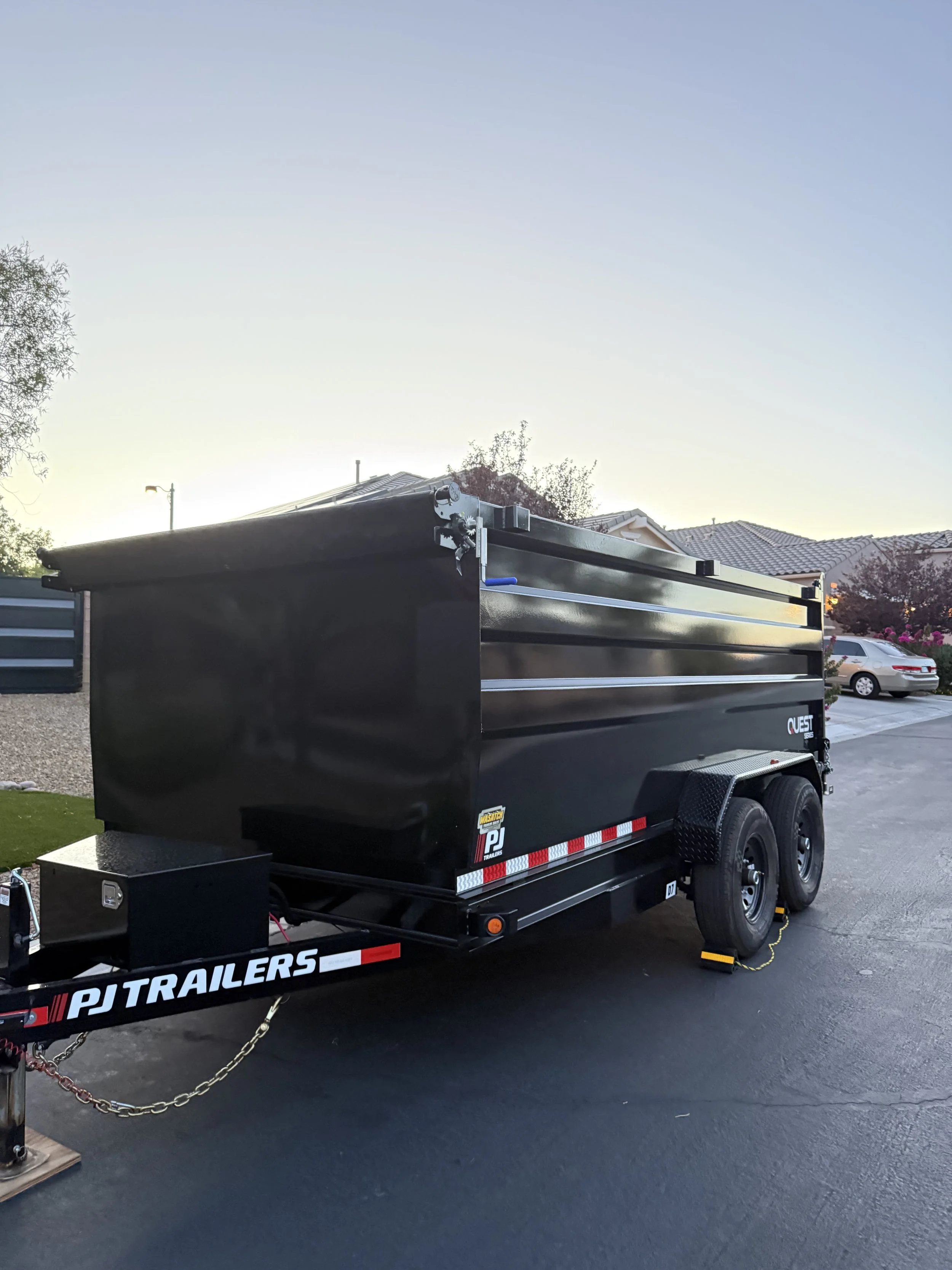 A black enclosed trailer with trailer wheels, hitch, and chains, parked on a driveway with houses and cars in the background.
