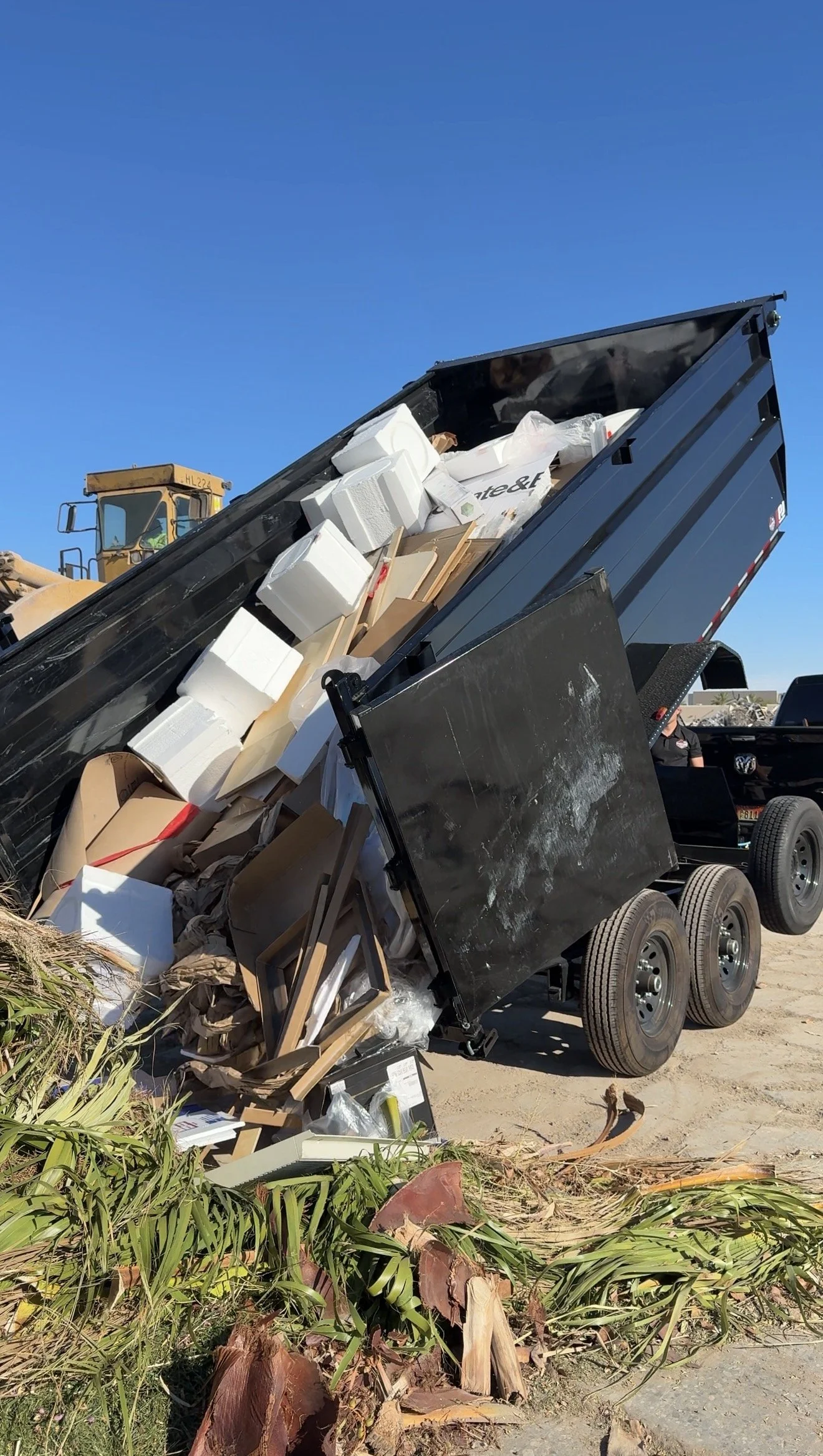 A black dump trailer tilted to unload debris, including cardboard boxes, foam packaging, picture frames, and plant materials, surrounded by green plants and dirt under a clear blue sky.