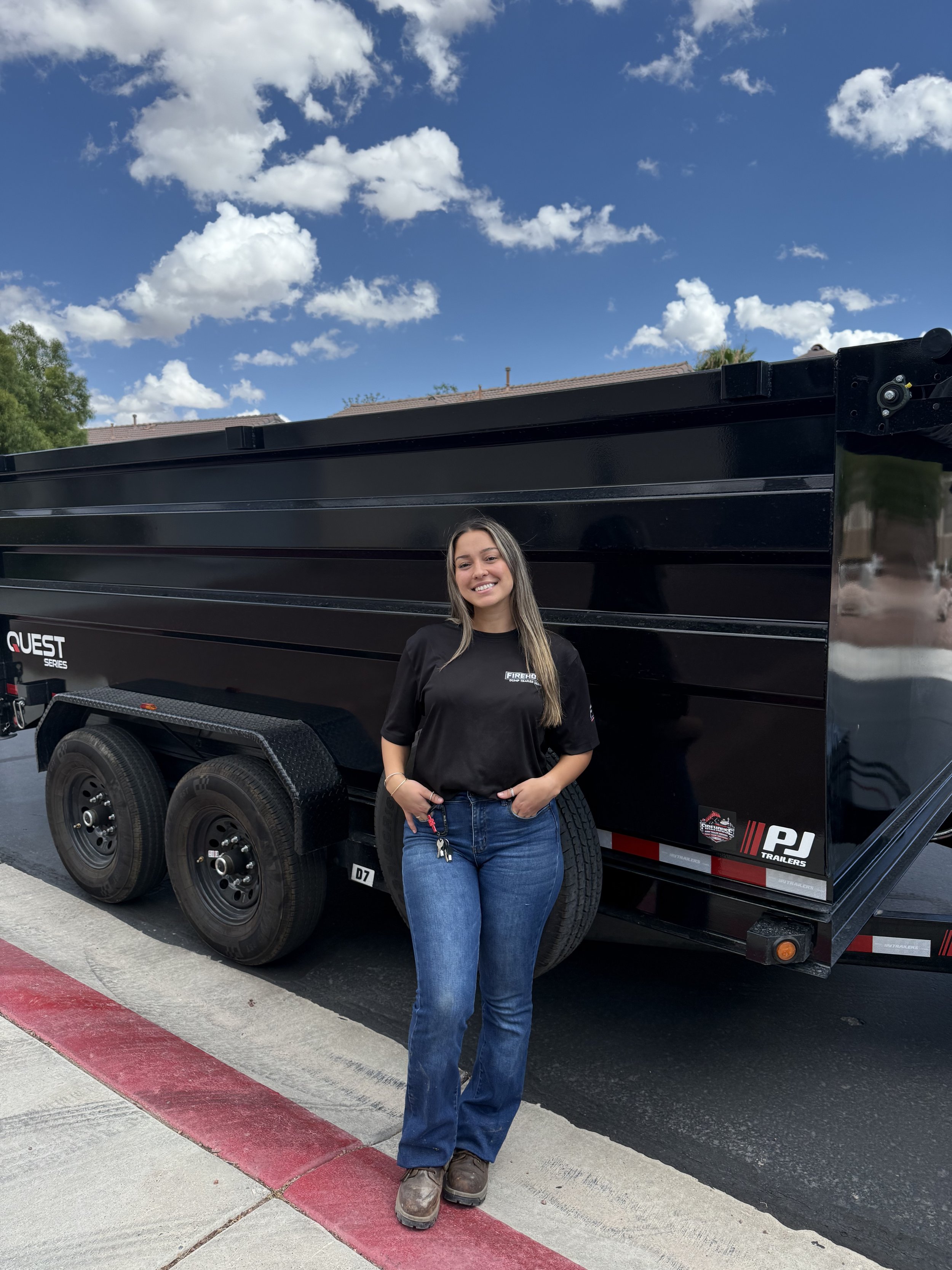 A young woman with long, dark blonde hair, wearing a black T-shirt and blue jeans, standing in front of a black trailer with large tires under a blue sky with scattered clouds.