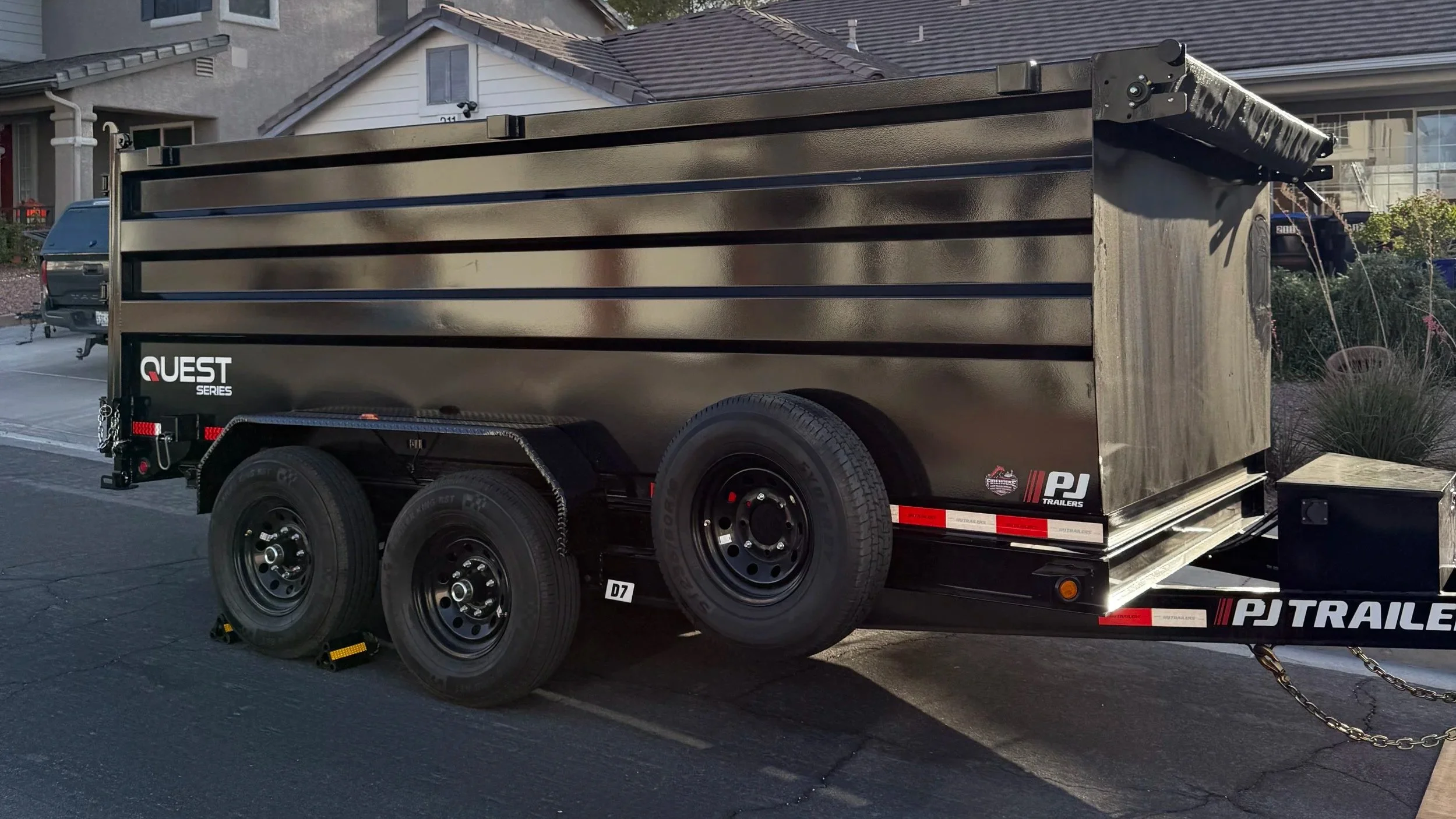 A black utility trailer with tandem wheels parked on a driveway in front of houses, with a small box secured on the front right corner.