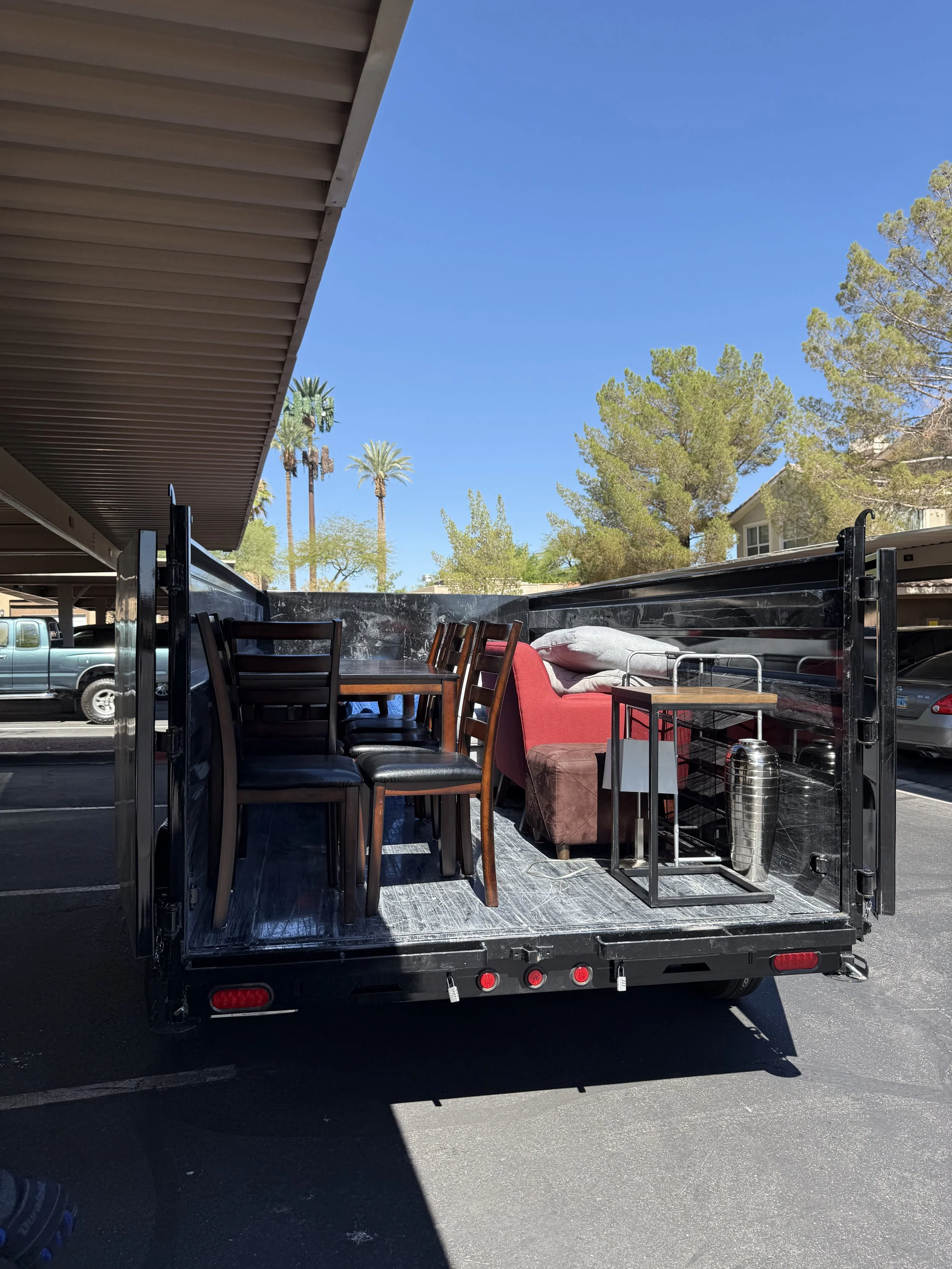 Back of a moving truck filled with furniture and household items parked in a lot under a building with a metal roof. View of blue sky and green trees in the background.