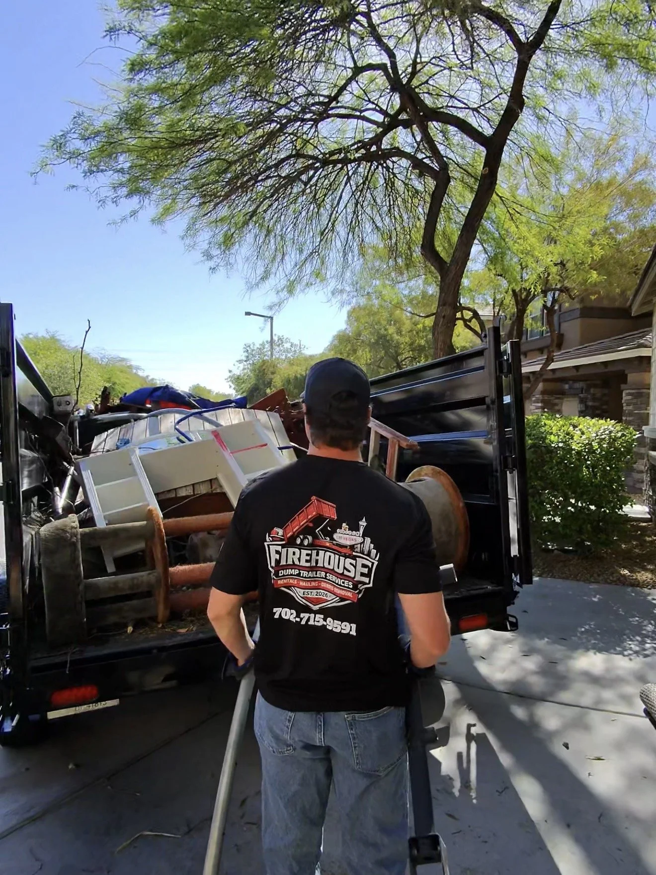Man wearing a black t-shirt with a firehouse logo and phone number on the back, standing in front of a loaded truck with furniture and household items, under a large tree on a clear sunny day.