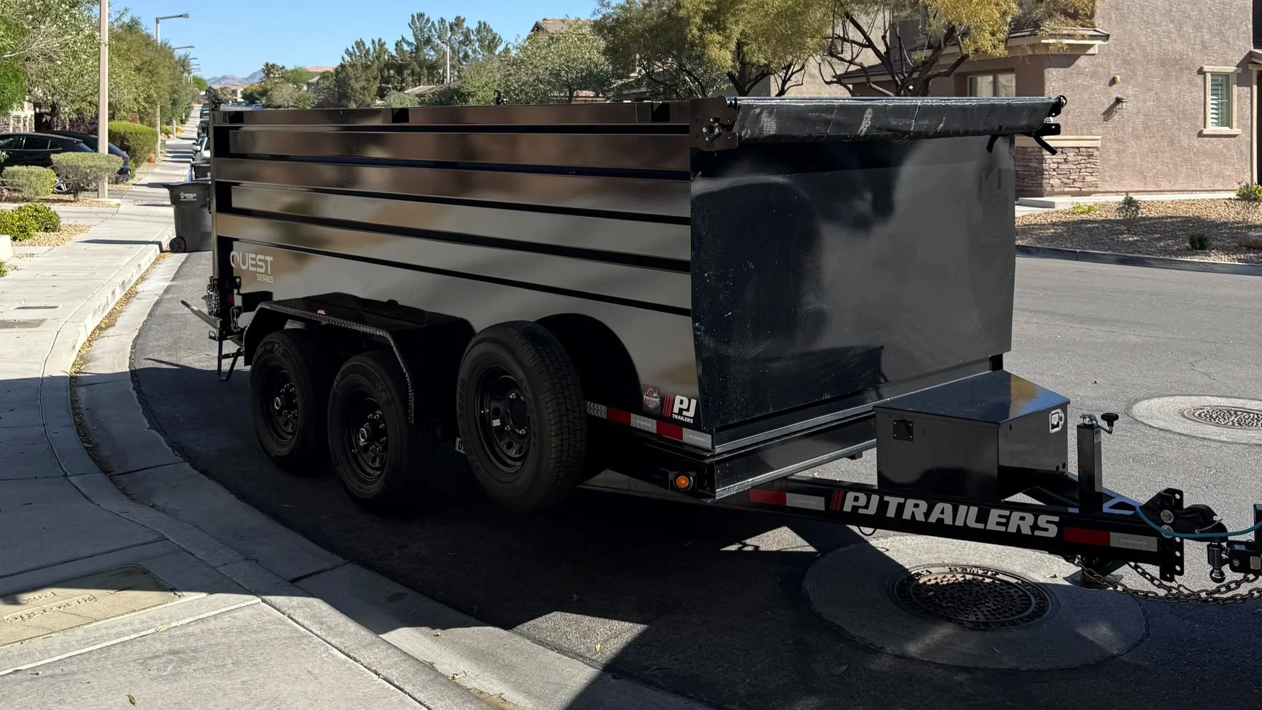Black enclosed utility trailer parked on a residential street next to a curb, with trees and houses in the background.
