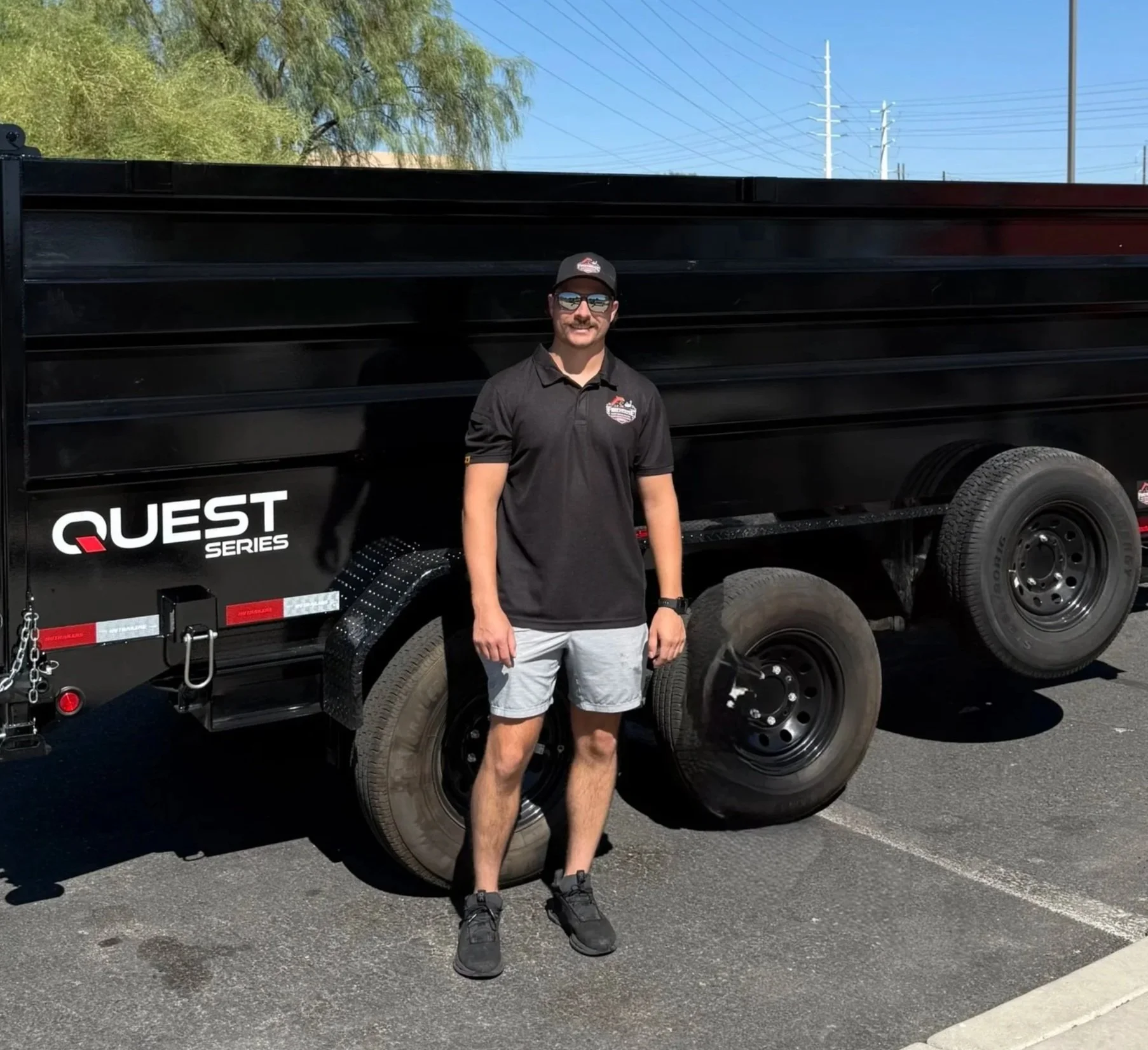 A man standing in front of a black utility trailer with the logo 'Quest Series' on the side. He is wearing a black polo shirt with a logo on the left side, gray shorts, black sneakers, a black cap, and sunglasses. The background features a clear blue sky, some green trees, and utility poles.