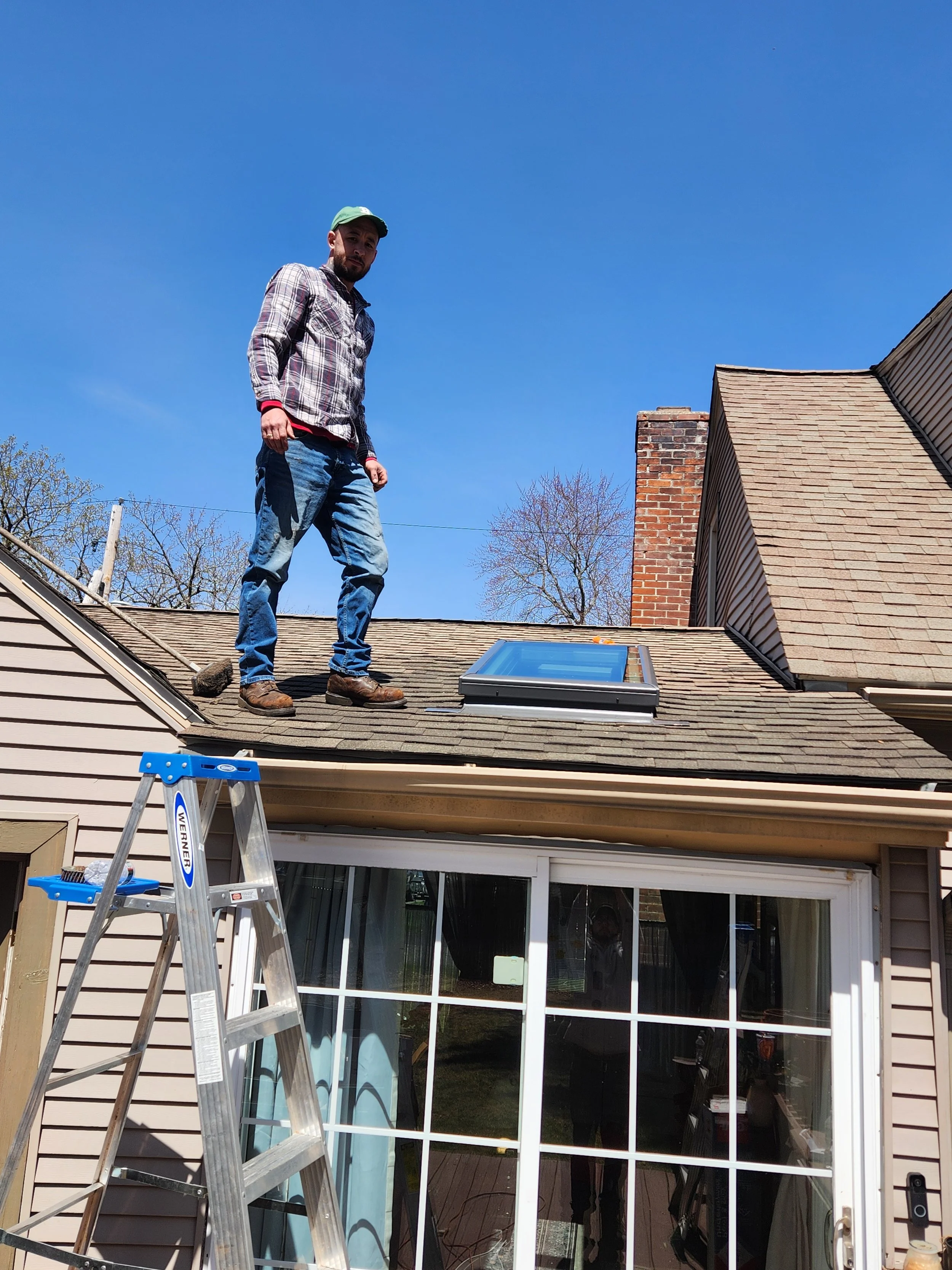 A man standing on a roof next to a skylight window with a ladder nearby, in front of a house with beige siding and a sliding glass door, under a clear blue sky.