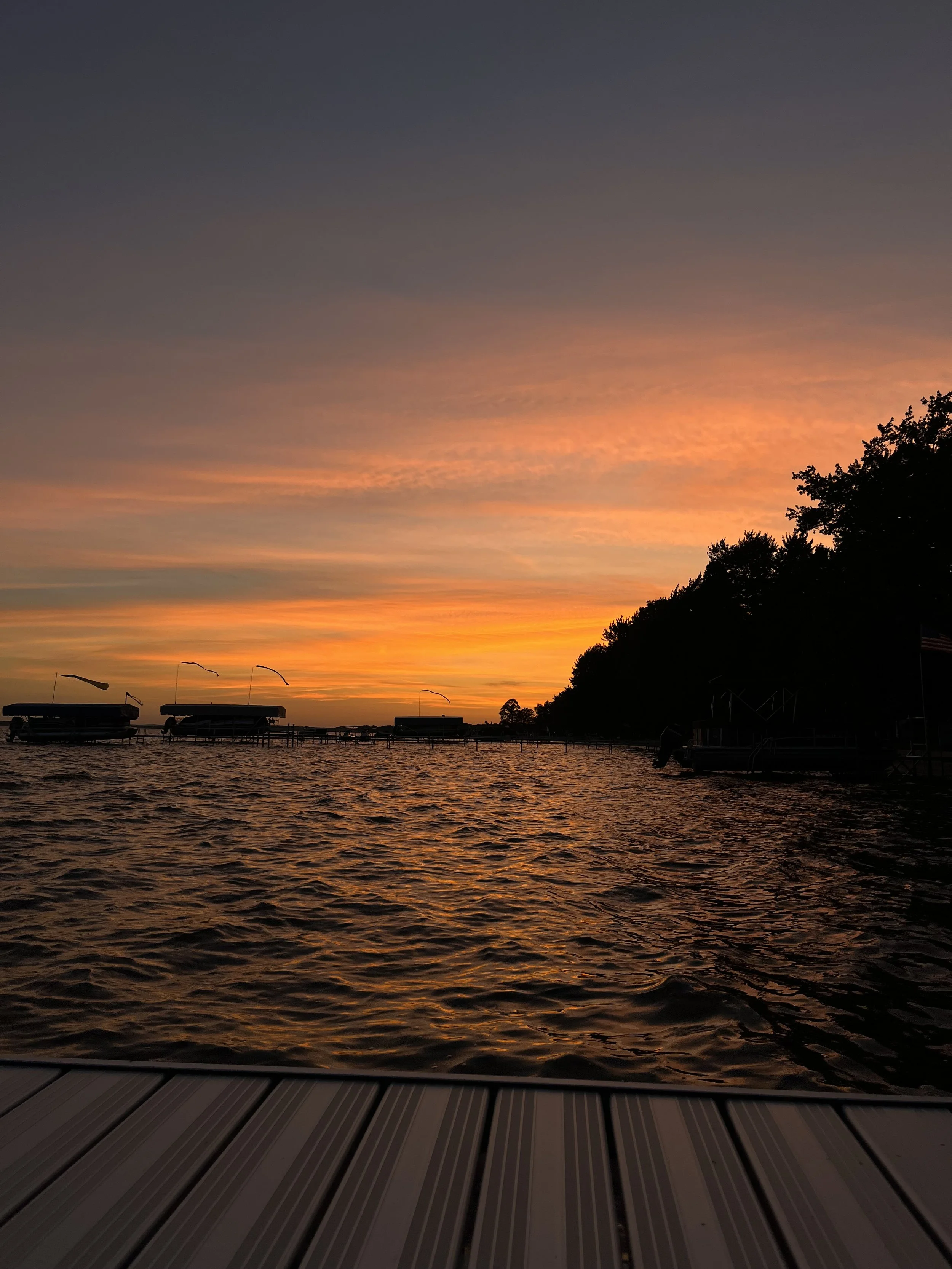 Sunset over a lake with boats anchored at a pier and a wooden dock in the foreground, silhouette of trees on the right.