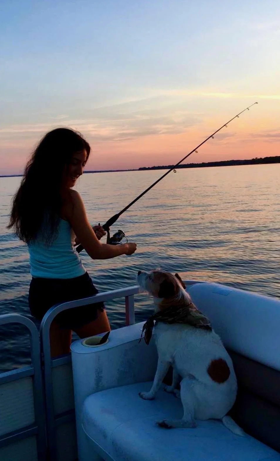 A woman fishing on a boat during sunset with a dog sitting on a cushioned seat next to her.