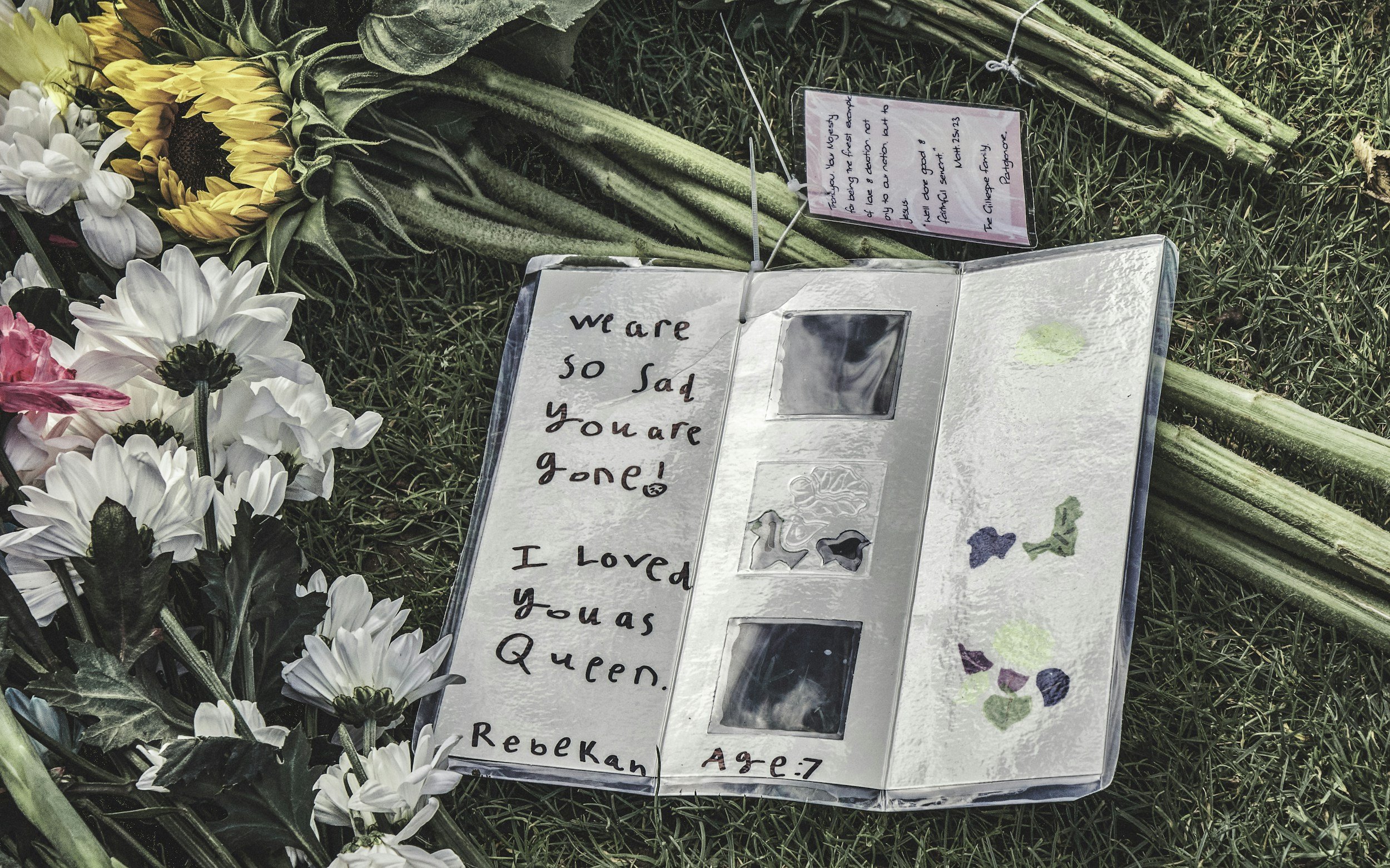 A memorial with flowers and a handwritten note that reads: "We are so sad you are gone! I loved you as Queen. Rebekah, Age 7." The memorial includes photos and heartfelt messages, placed on grass.