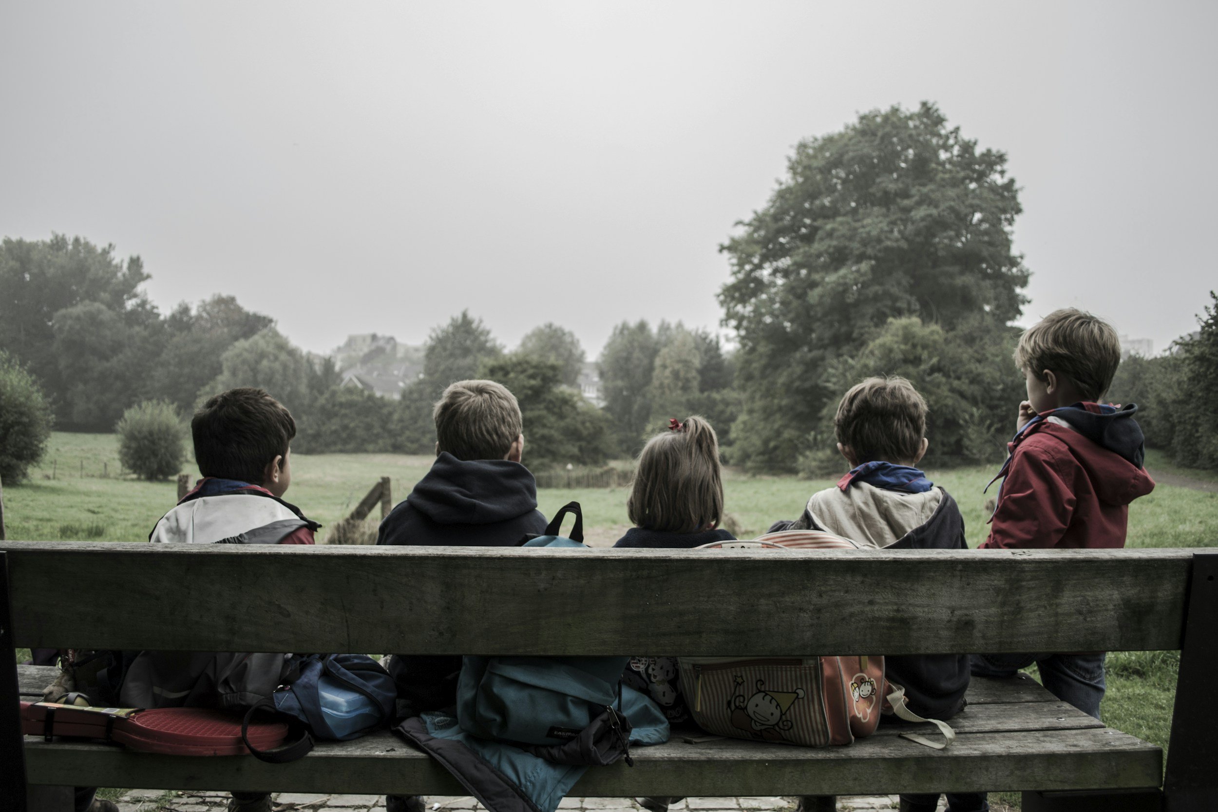 Five children sitting on a park bench looking at a green field and trees under a cloudy sky.