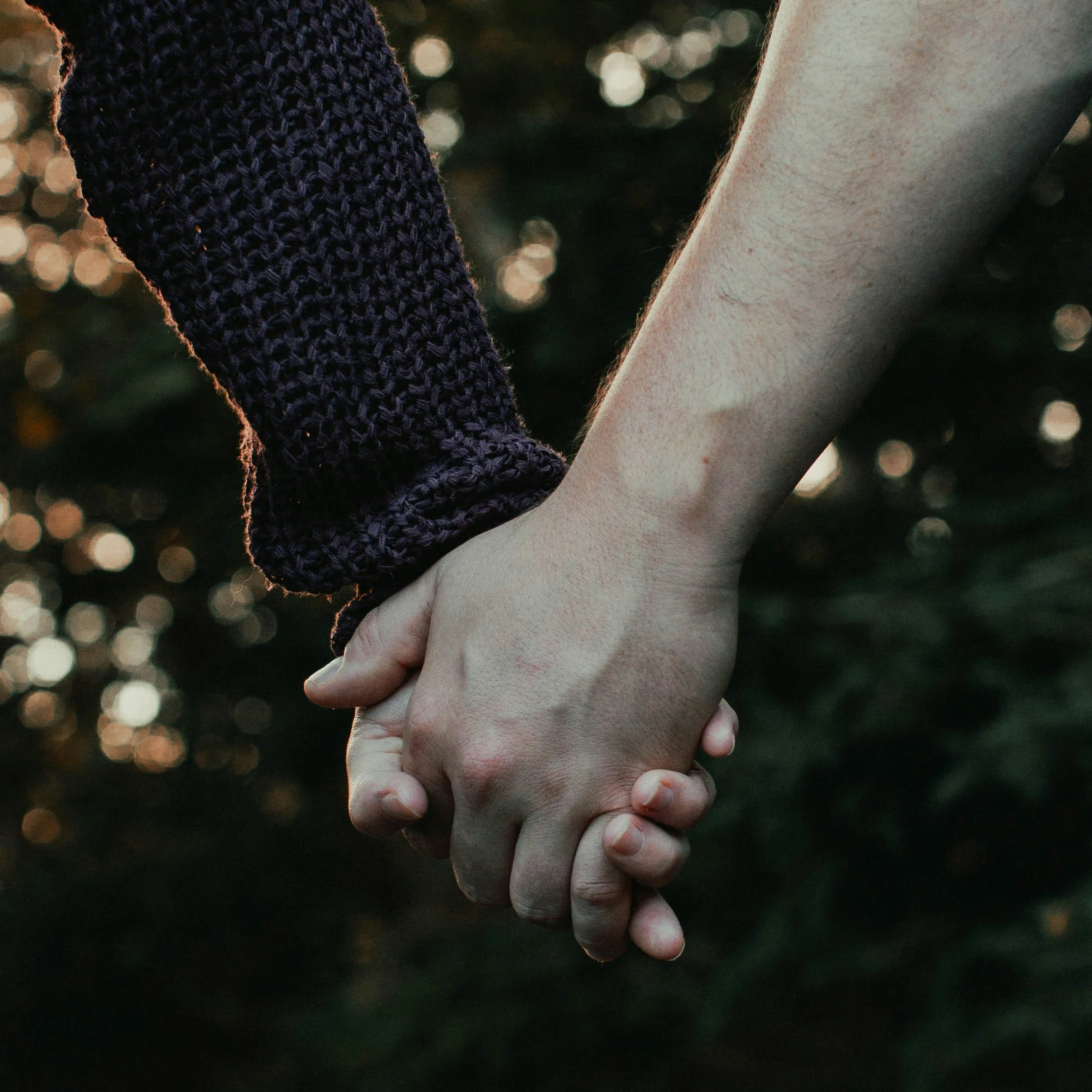 Two hands, one with a dark knit sleeve, clasped together with a gentle grip against a blurred background of bokeh lights.