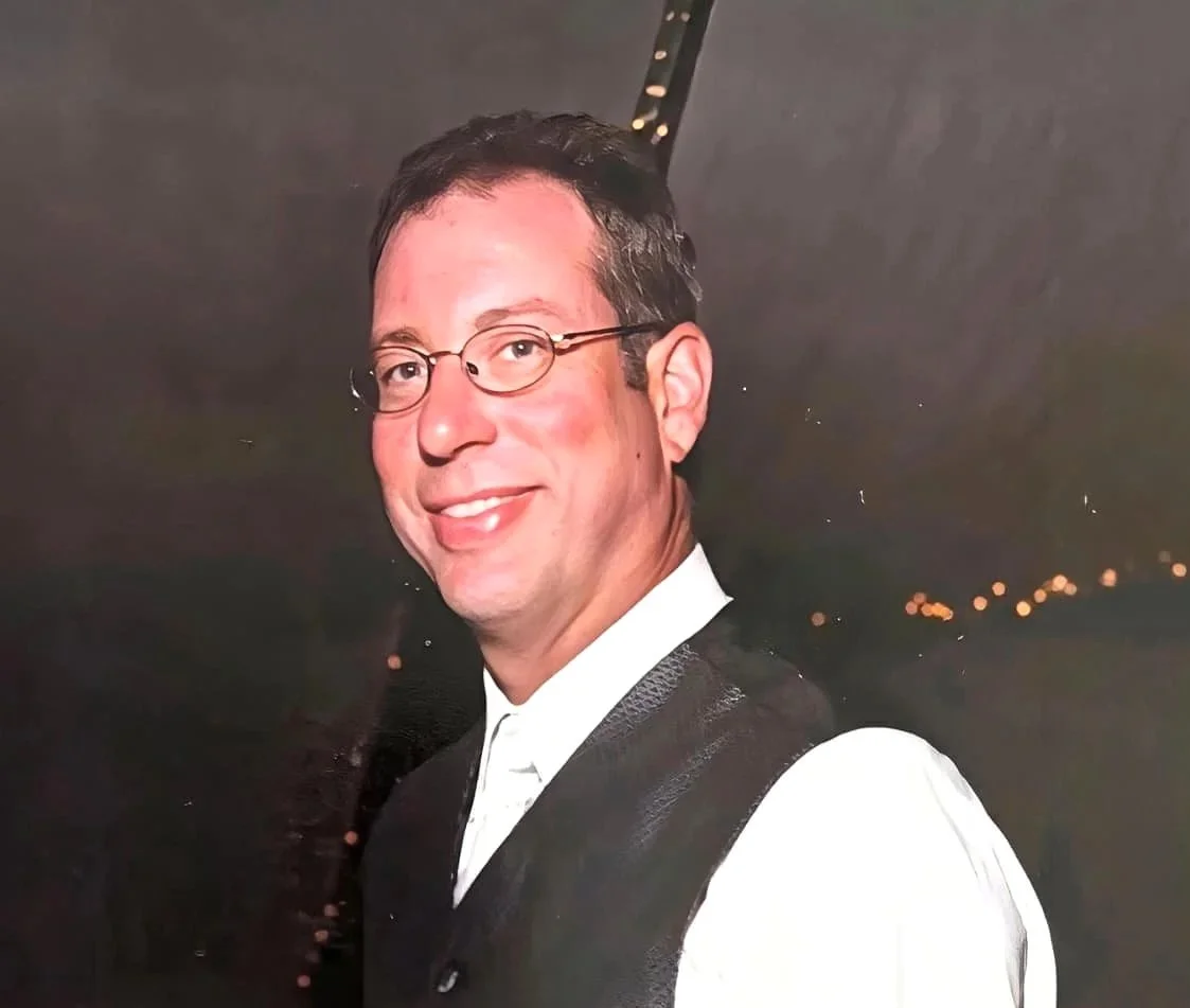 A smiling man with short dark hair, glasses, wearing a white shirt and black vest, at night with a dark background and lights.
