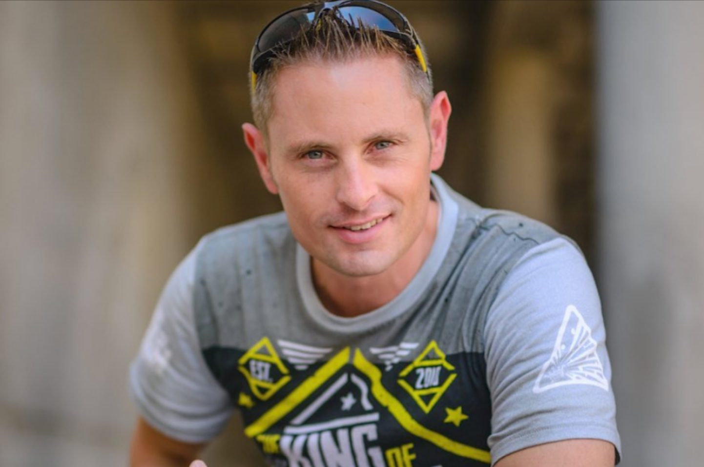 A young man with short blond hair, light skin, and Blue eyes, wearing sunglasses on his head and a casual gray t-shirt with graphic designs, smiling outdoors.