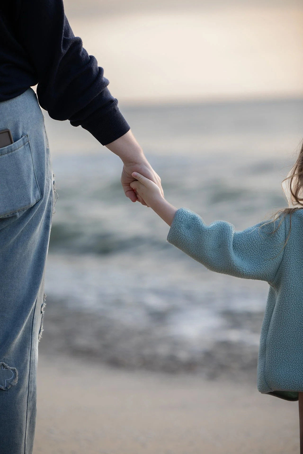 A person in a black sweater holding hands with a young girl in a blue sweater at the beach.