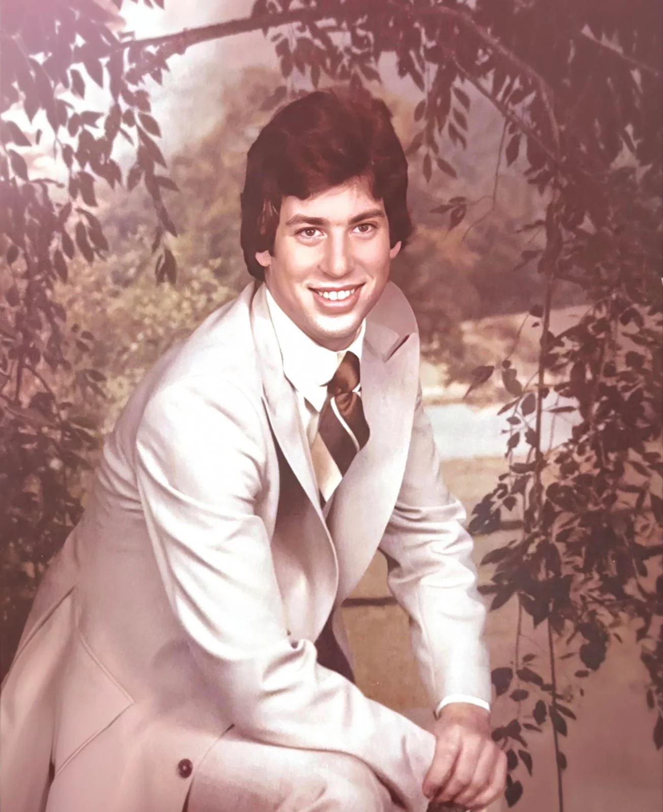 A man with dark hair, wearing a light-colored suit, white shirt, and striped tie, posing outdoors among trees and foliage, smiling at the camera.