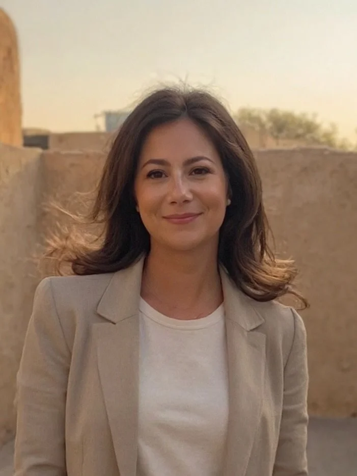 A woman with shoulder-length dark hair, wearing a cream-colored blazer and white shirt, standing outdoors during sunset with a textured beige wall in the background.