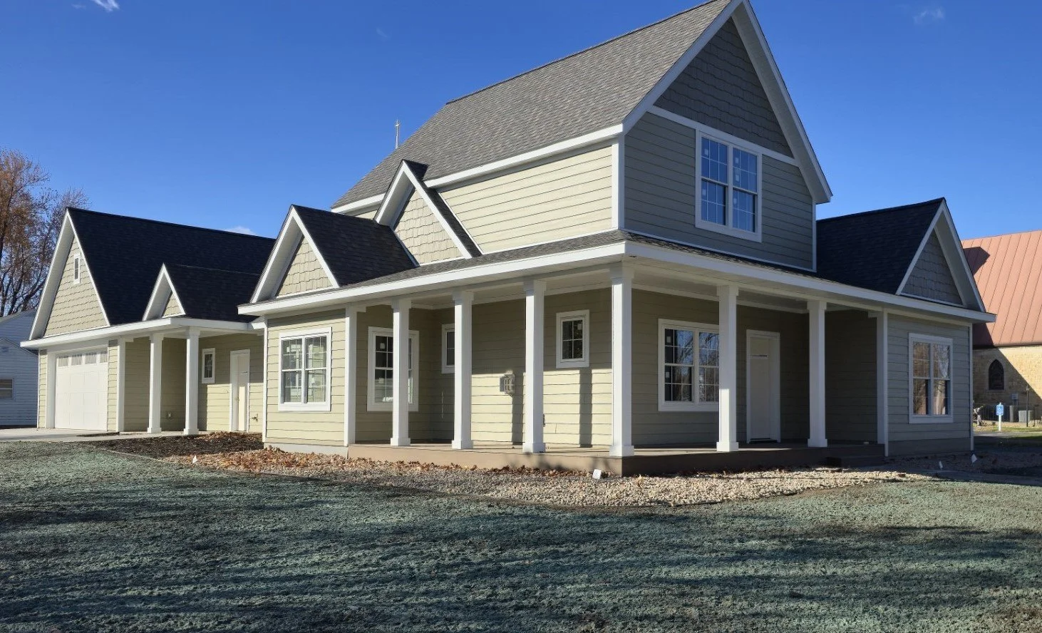 Newly constructed two-story suburban house with beige siding, white trim, and a covered front porch with columns, set against a clear blue sky.