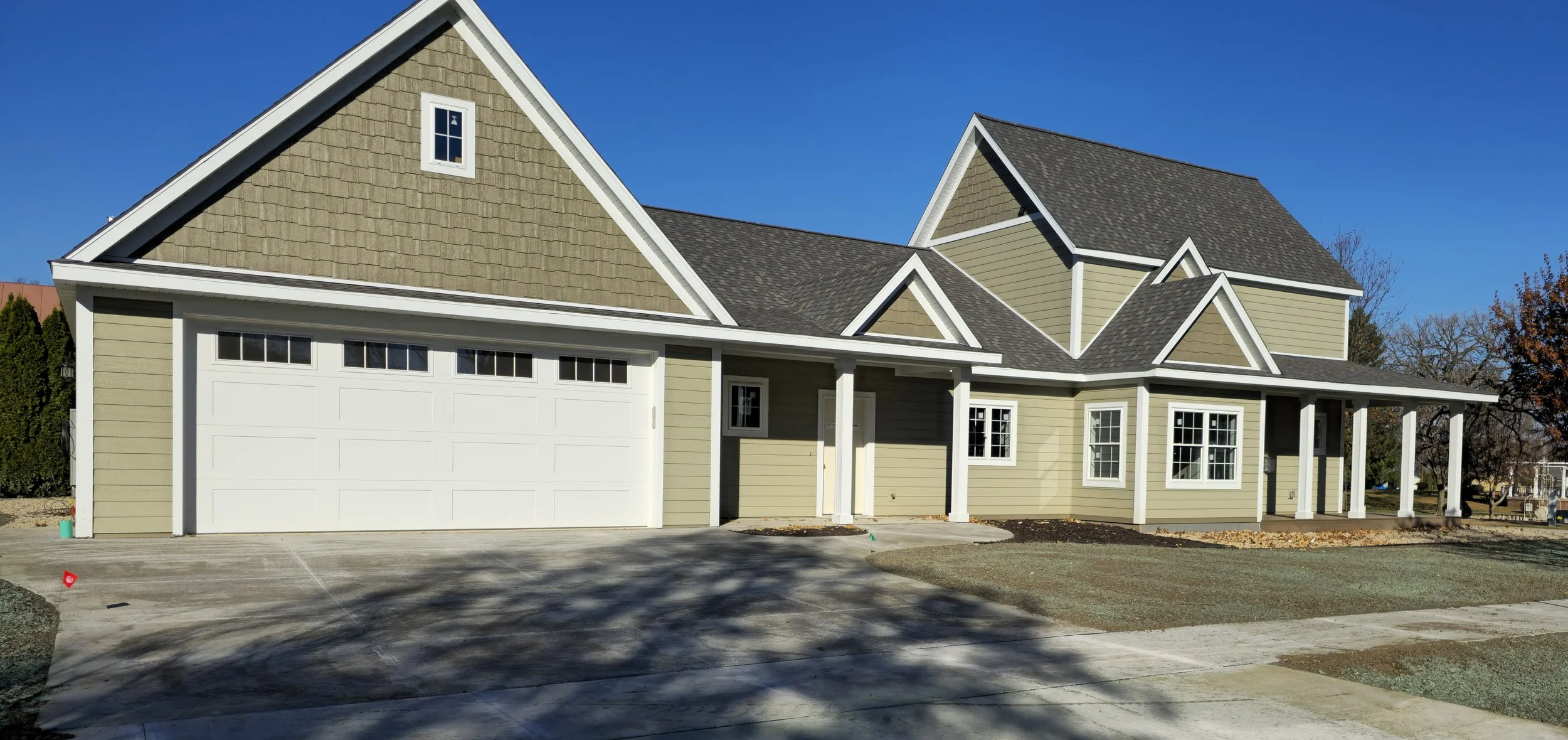 Newly constructed two-story house with a white garage door, multiple windows, and a front porch supported by white columns, with a driveway and front yard under construction.