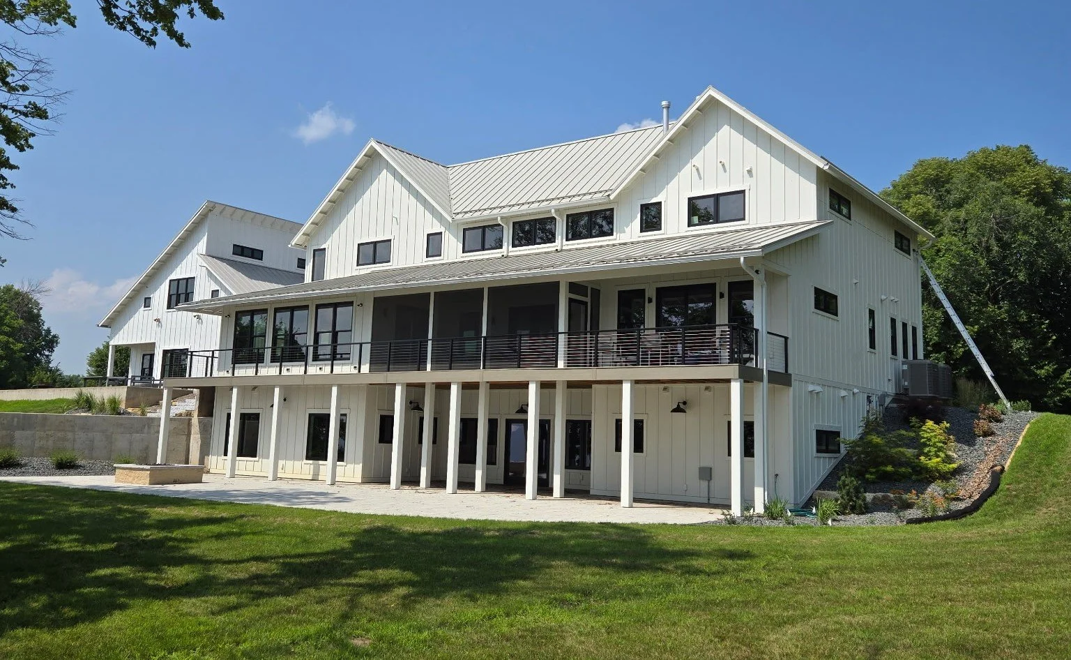 A modern white multi-story house with a metal roof, large windows, and a wrap-around porch, set against a clear blue sky and surrounded by green lawn and trees.