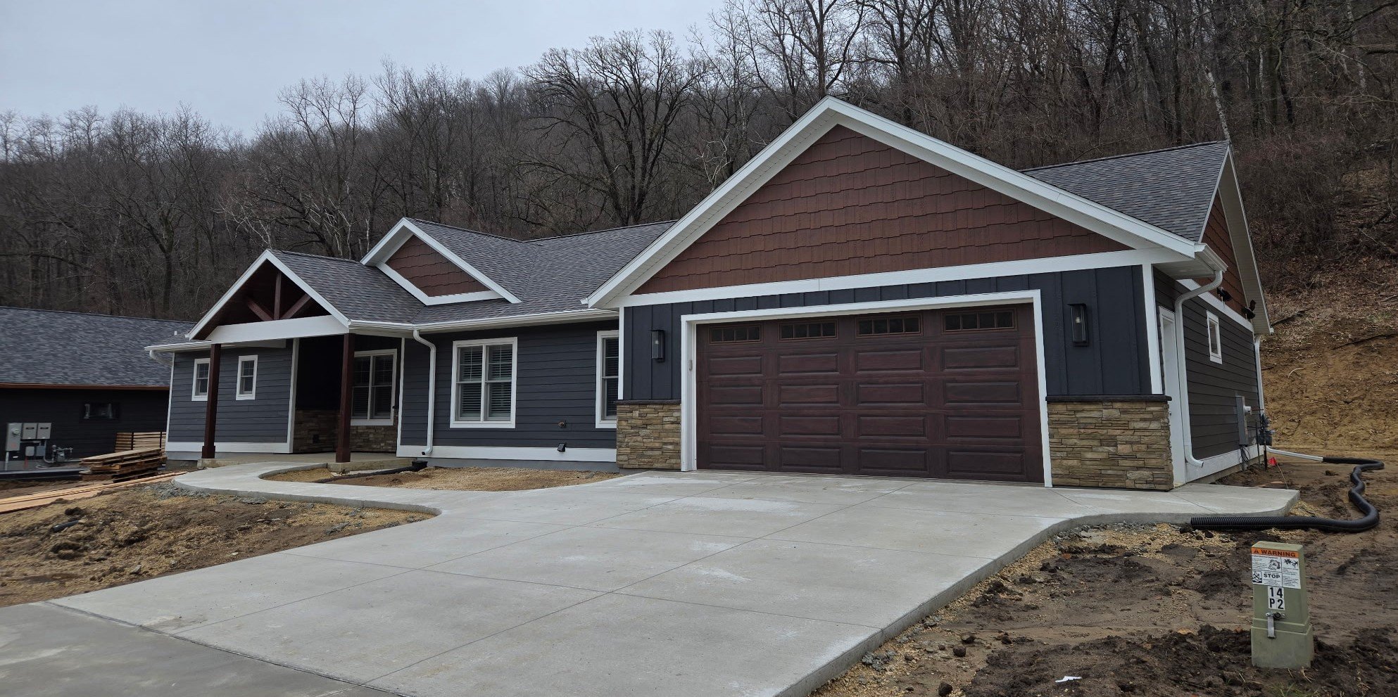 Newly constructed house with a dark blue exterior, a stone accent on the lower part of the garage, and a gray shingle roof. There is a concrete driveway and a small covered porch at the front, with some landscaping in progress around it. The background shows leafless trees on a hillside during late fall or winter.