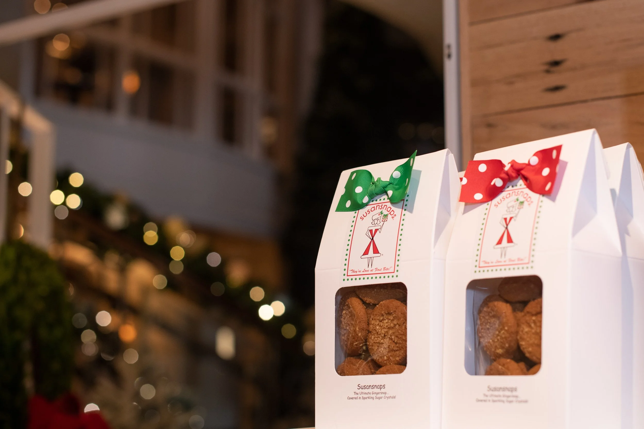 Boxed cookies labeled Susansnaps with red and green bows, with a festive background of blurred Christmas lights and decorations.