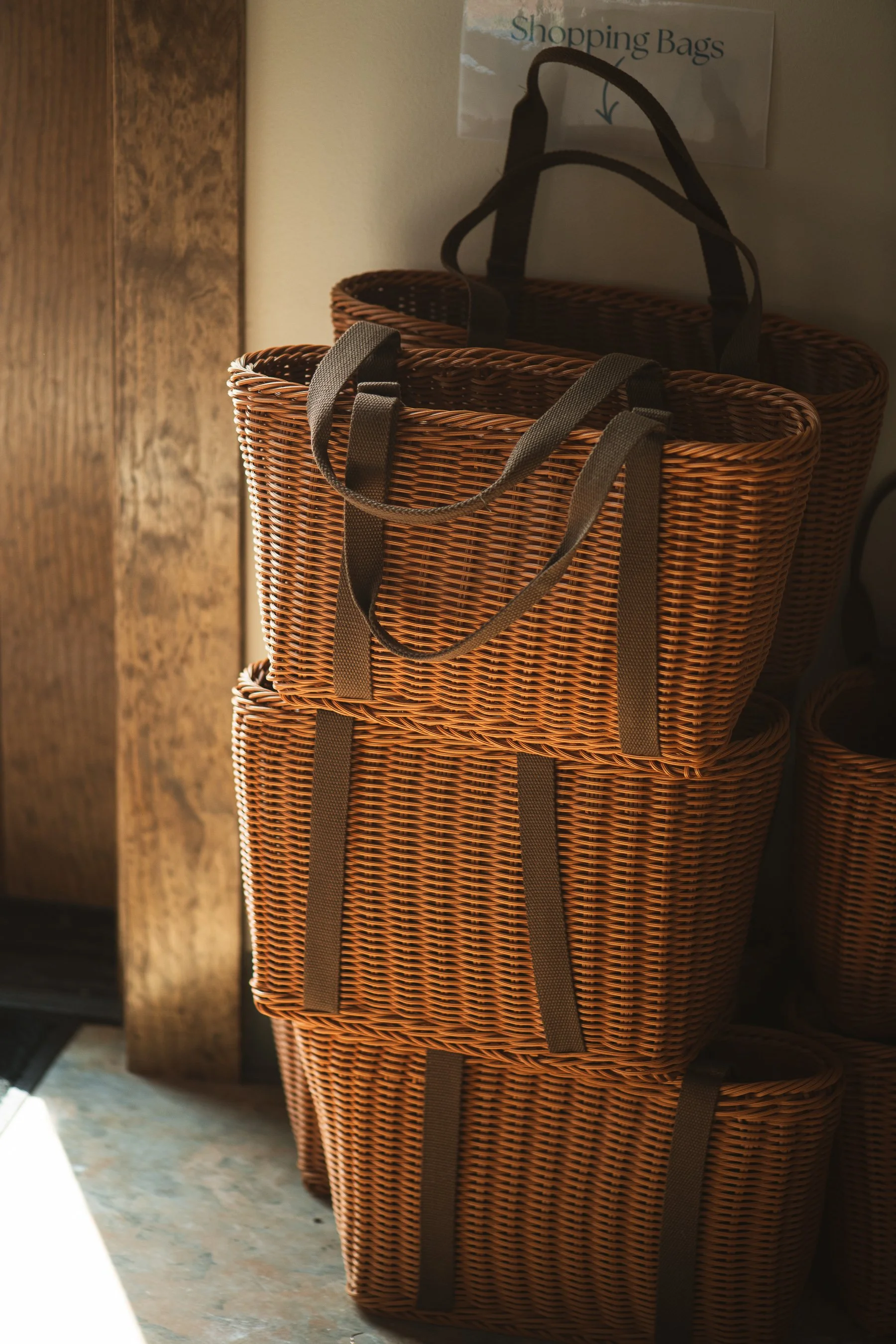 Three stacked wicker shopping baskets with dark handles, placed next to a wooden wall with a sign that reads 'Shopping Bags' and an arrow pointing downward.