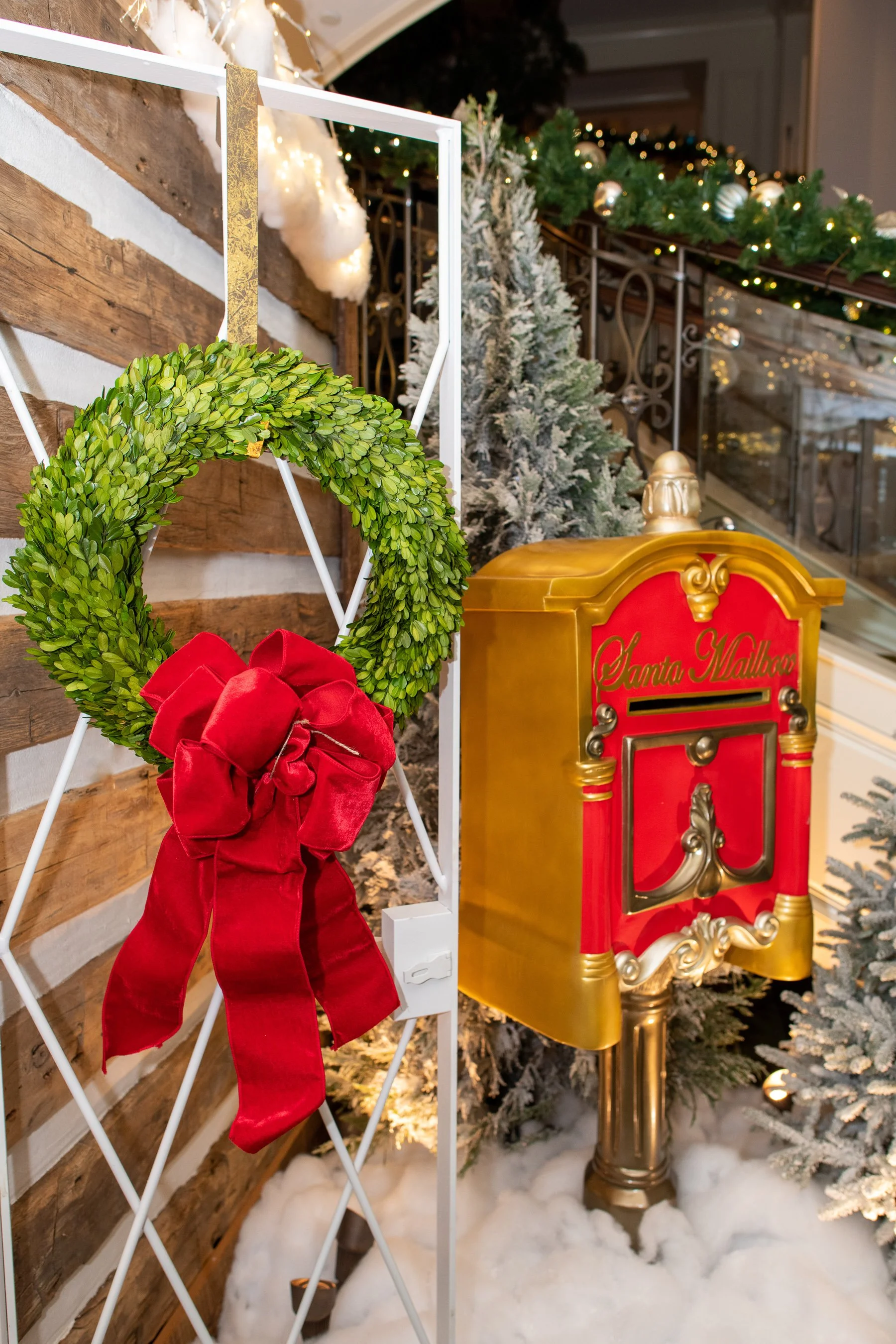 Decorated Christmas scene with a green wreath and red bow, a yellow and red Santa mailbox, snow-covered ground, and Christmas trees in the background with Christmas ornaments and lights.