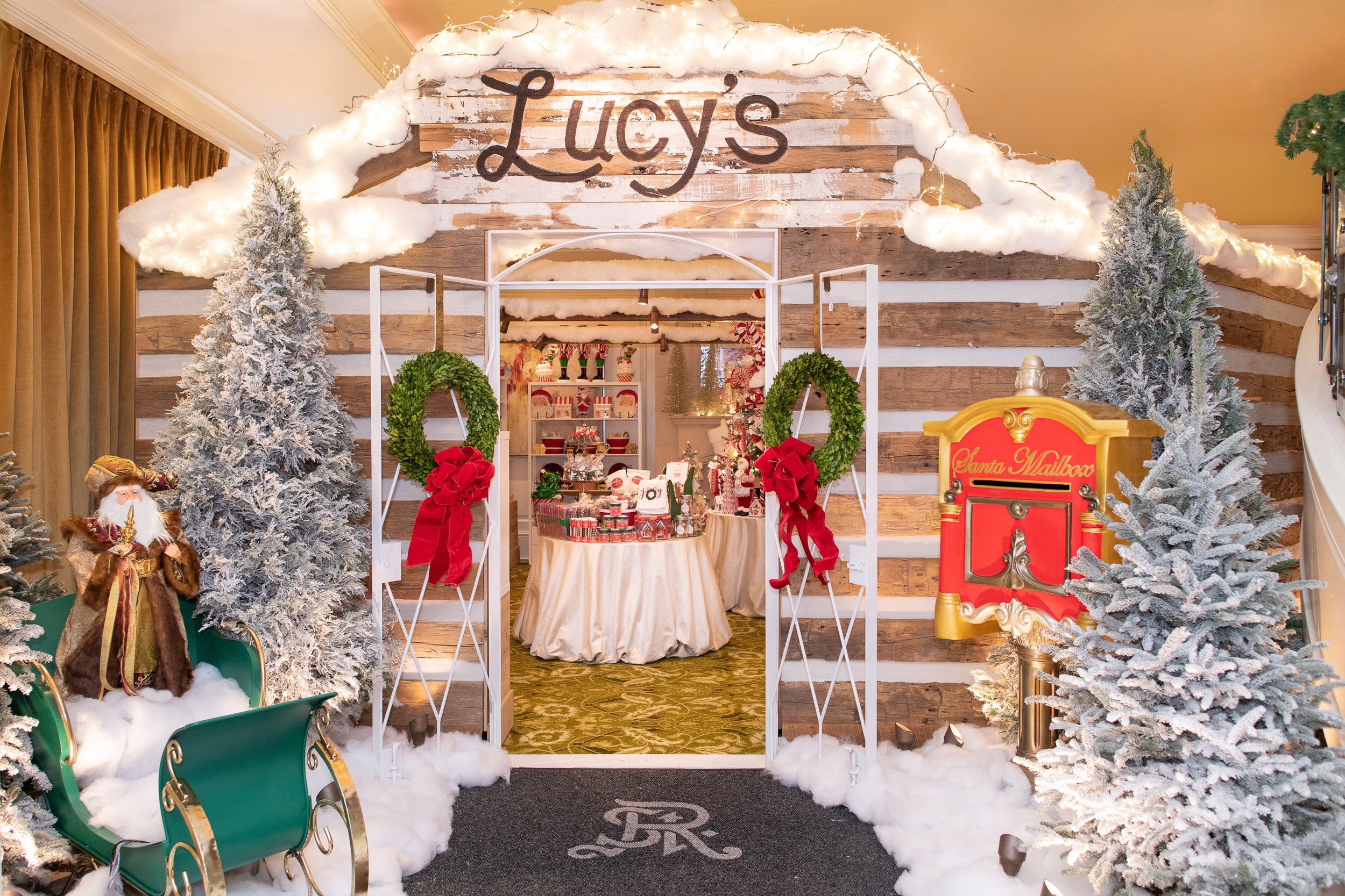 St. Regis Atlanta Astor Chalet with snowy trees, a gingerbread man figure, a red mailbox labeled 'Santa Mailbox', and a display of holiday treats and decorations in a rustic setting with wooden accents and string lights.